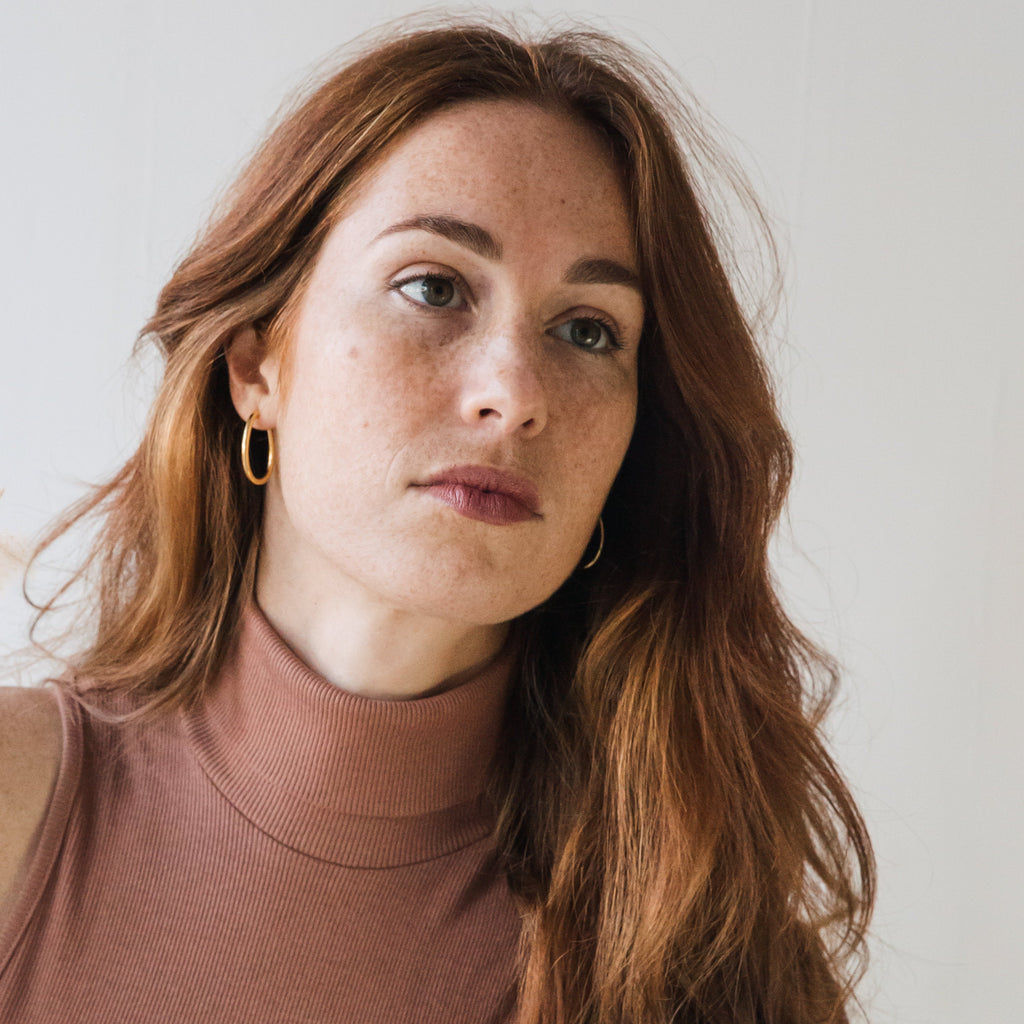 A woman with long red hair wears Anine Oval Hoops, gazing slightly to the side against a plain background.