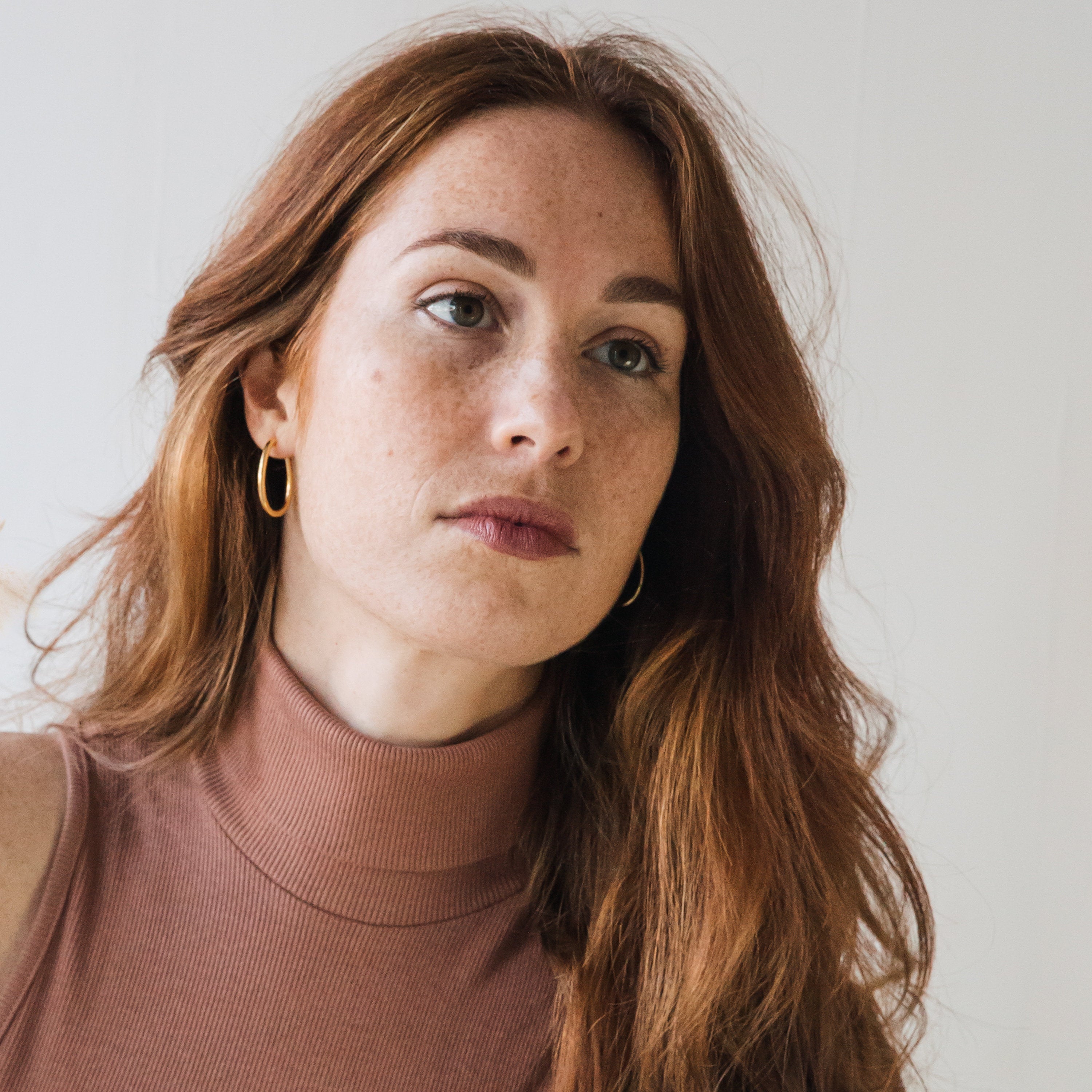 A woman with long red hair wears Anine Oval Hoops, gazing slightly to the side against a plain background.