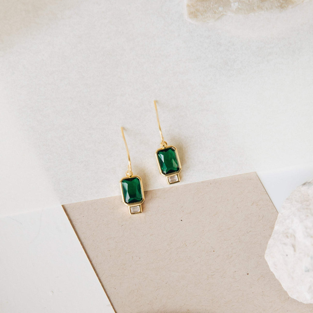 The Emerald Drop Earrings feature rectangular green stones and small clear accents, showcased against a neutral background.