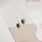 The Emerald Drop Earrings feature rectangular green stones and small clear accents, showcased against a neutral background.