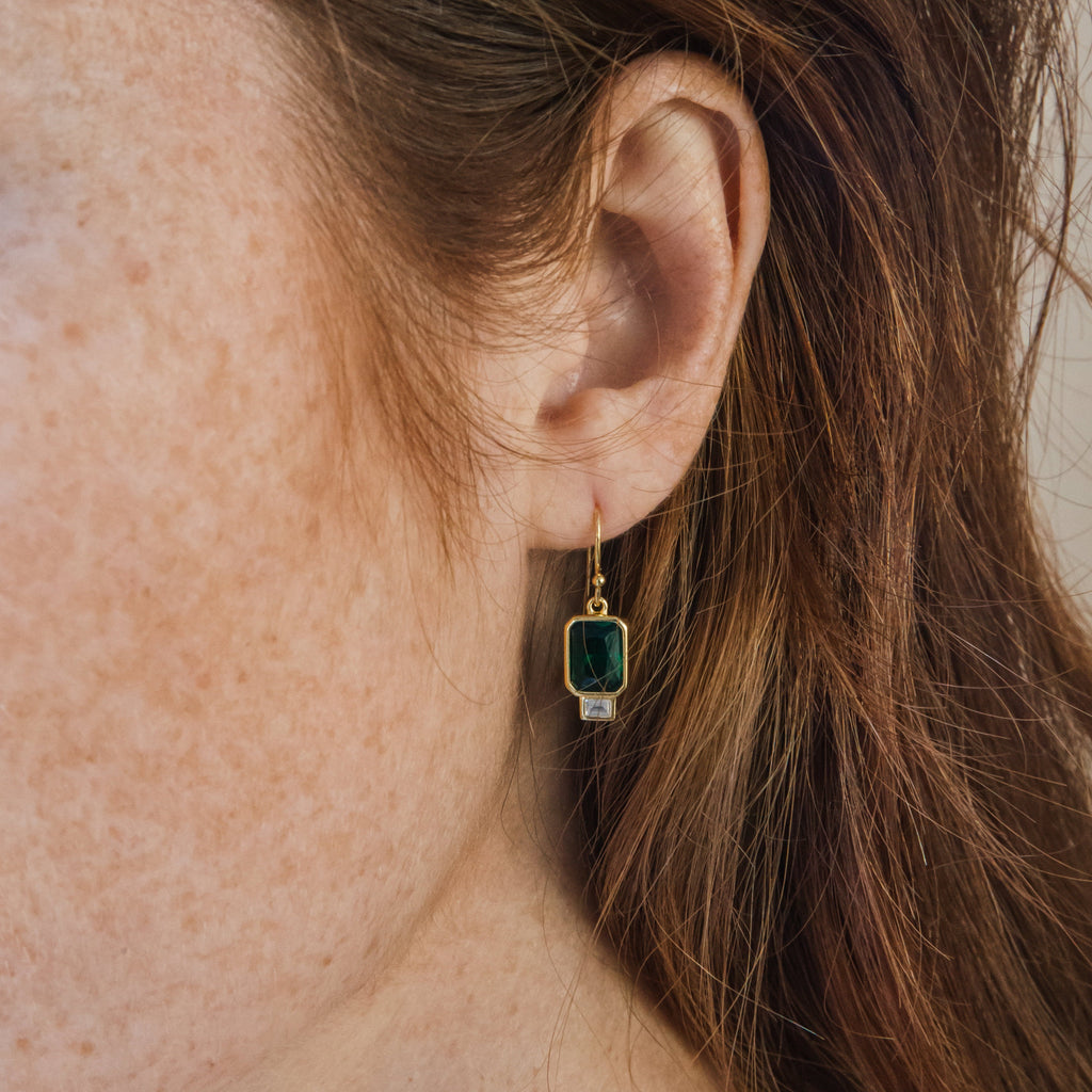 Close-up of a woman's ear wearing Emerald Drop Earrings, featuring teardrop-shaped dark green stones, with her brown hair gently framing the scene.