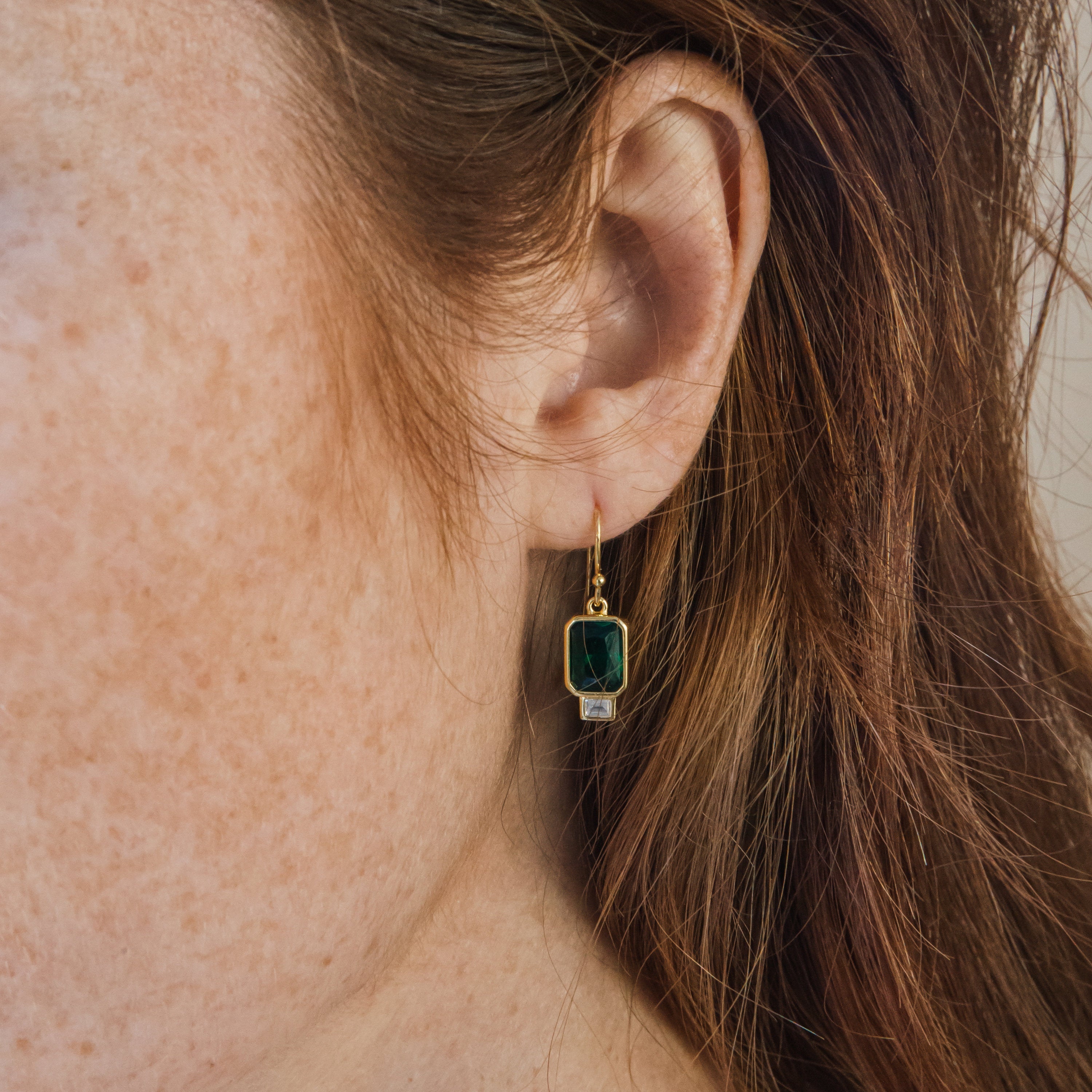 Close-up of a woman's ear wearing Emerald Drop Earrings, featuring teardrop-shaped dark green stones, with her brown hair gently framing the scene.