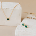 Malachite Studs and a gold necklace with geometric green stones are elegantly displayed on white textured blocks, accented by dried foliage. The malachite studs add a modern touch to this striking jewelry set.
