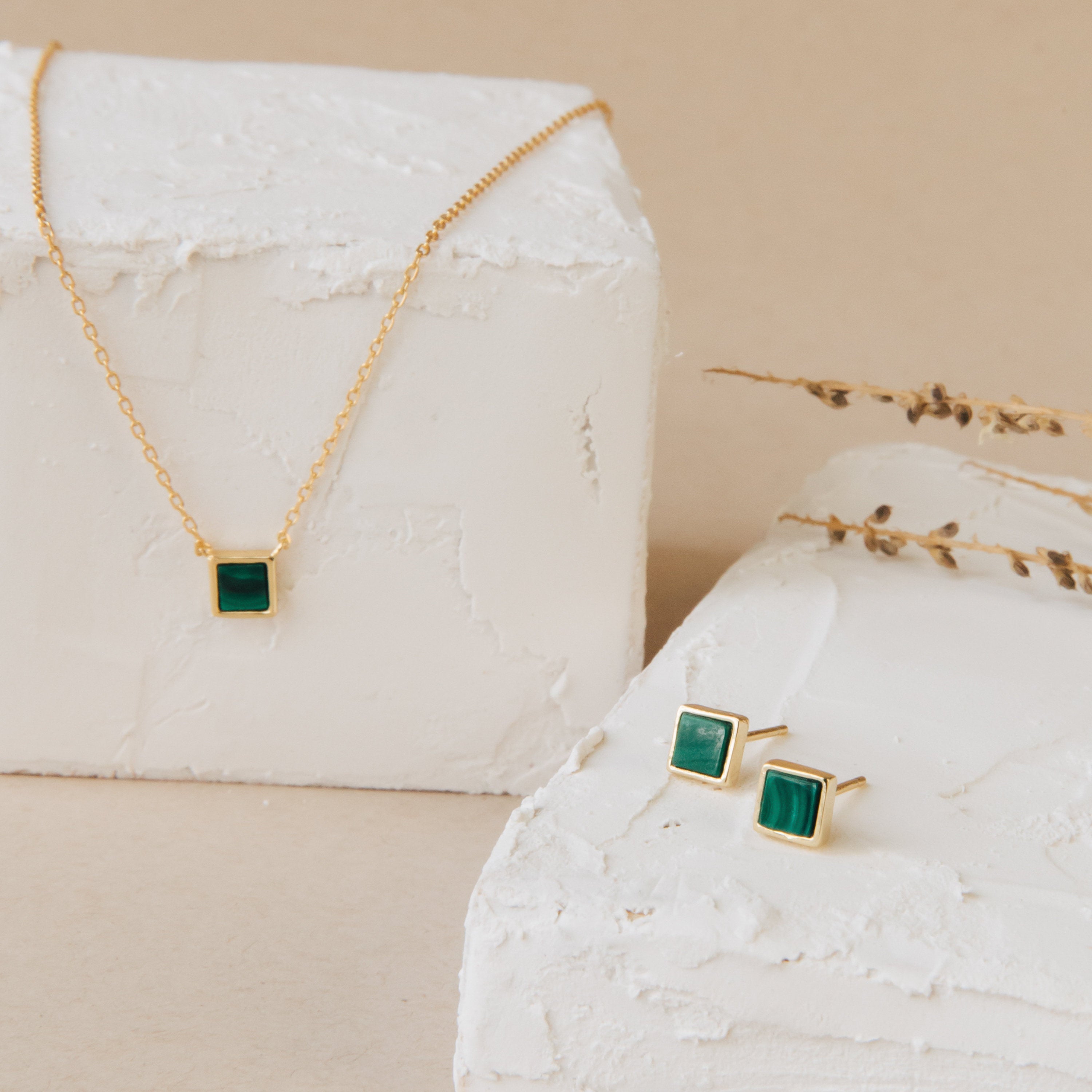 Malachite Studs and a gold necklace with geometric green stones are elegantly displayed on white textured blocks, accented by dried foliage. The malachite studs add a modern touch to this striking jewelry set.