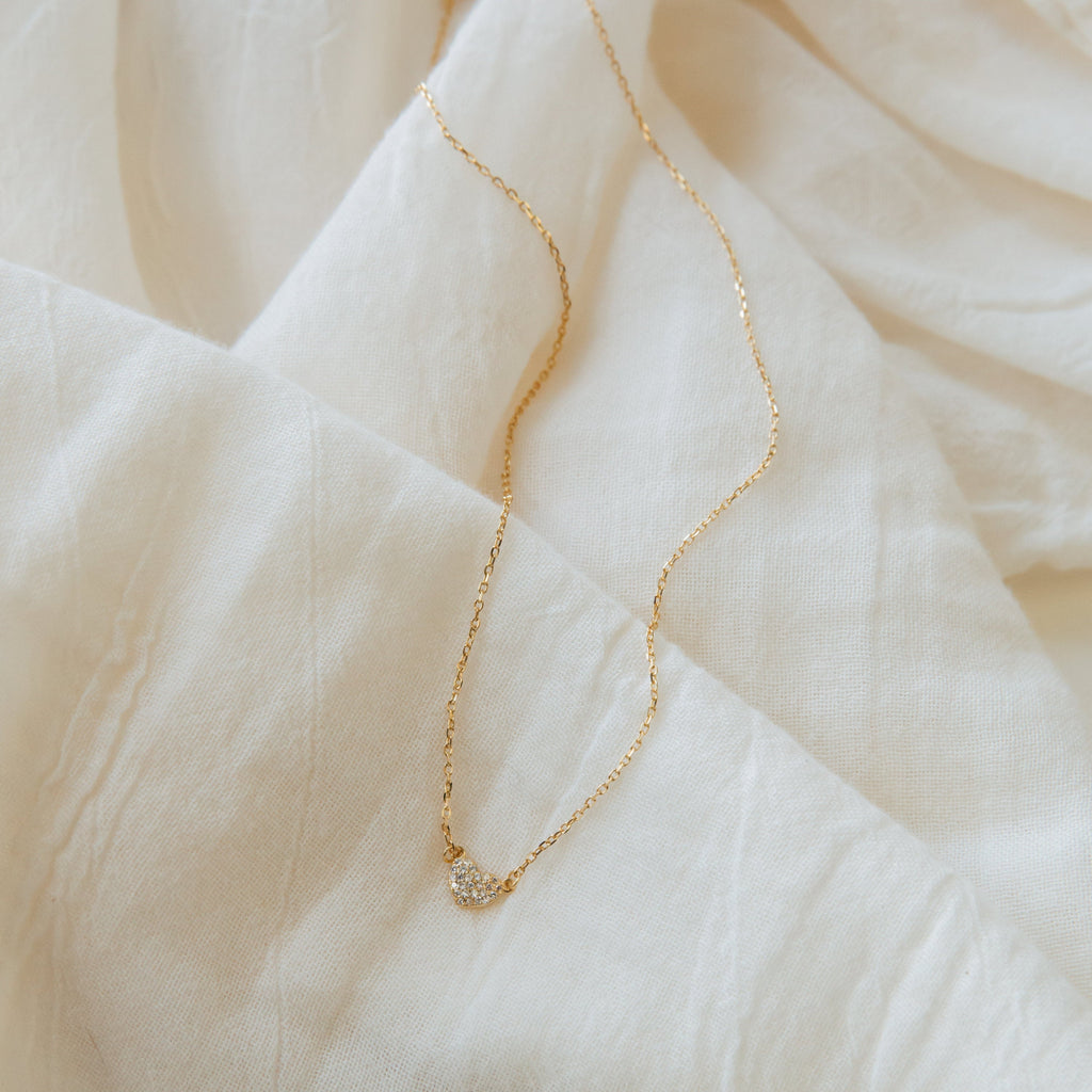 The Pave Heart Necklace from the Coquette Collection is a delicate gold chain with a small pave heart charm, displayed on soft, light fabric with gentle folds and texture.