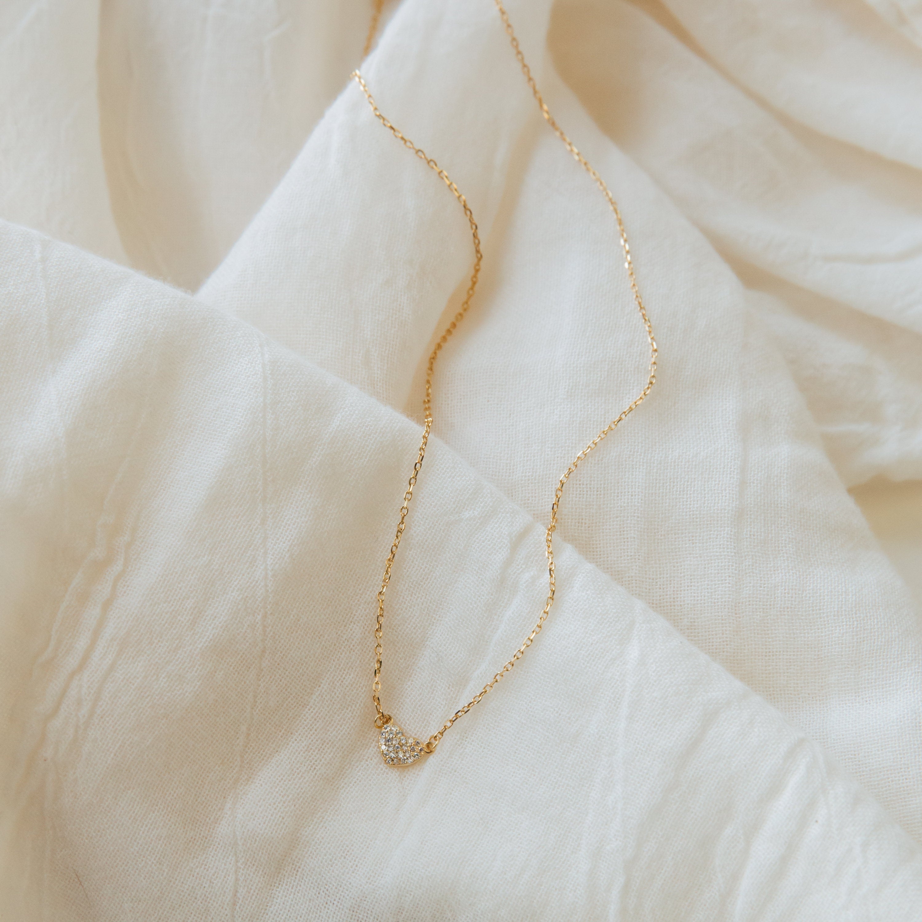 The Pave Heart Necklace from the Coquette Collection is a delicate gold chain with a small pave heart charm, displayed on soft, light fabric with gentle folds and texture.