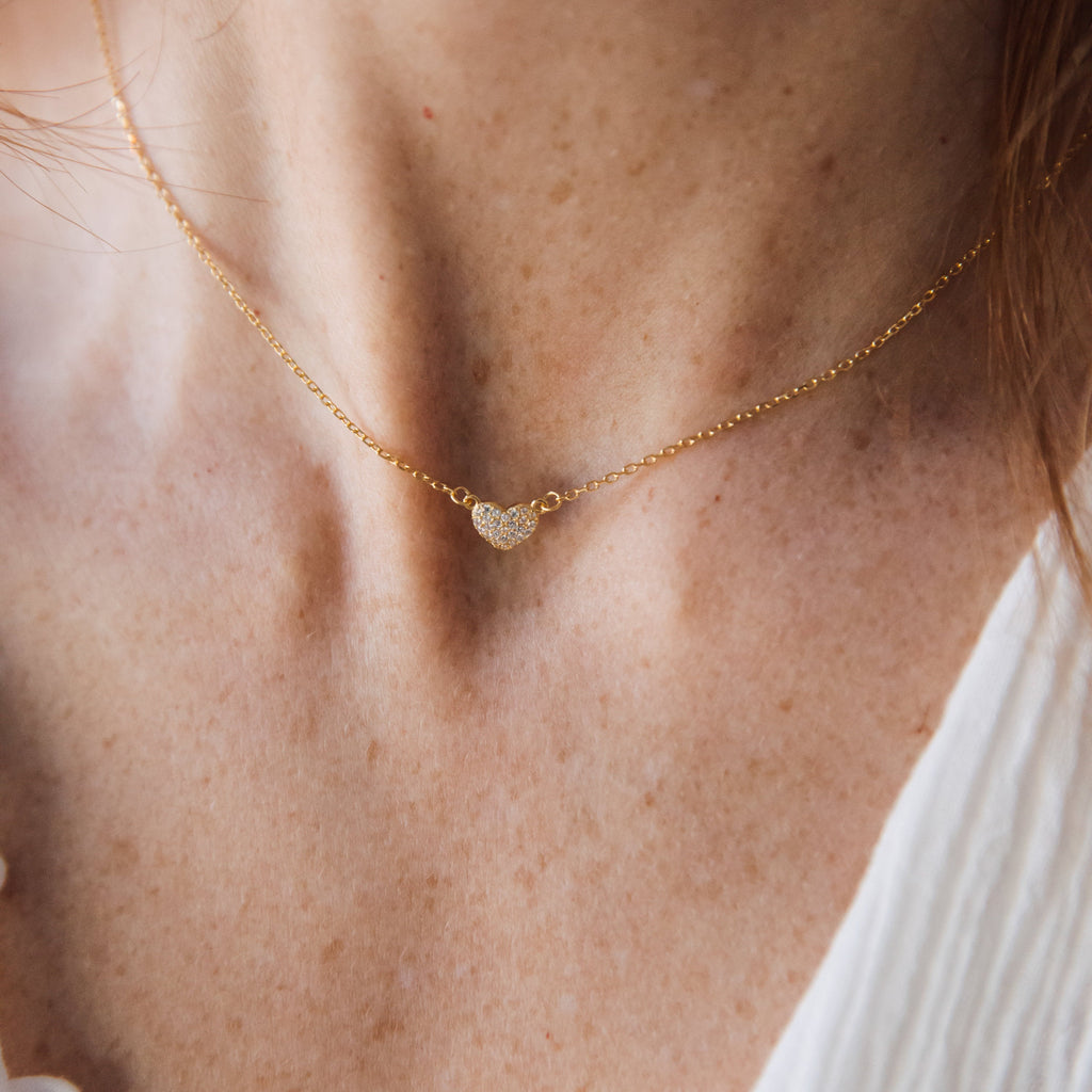 A minimalist Pave Heart Necklace rests on a gold chain, elegantly paired with a white top for an everyday look inspired by timeless style.
