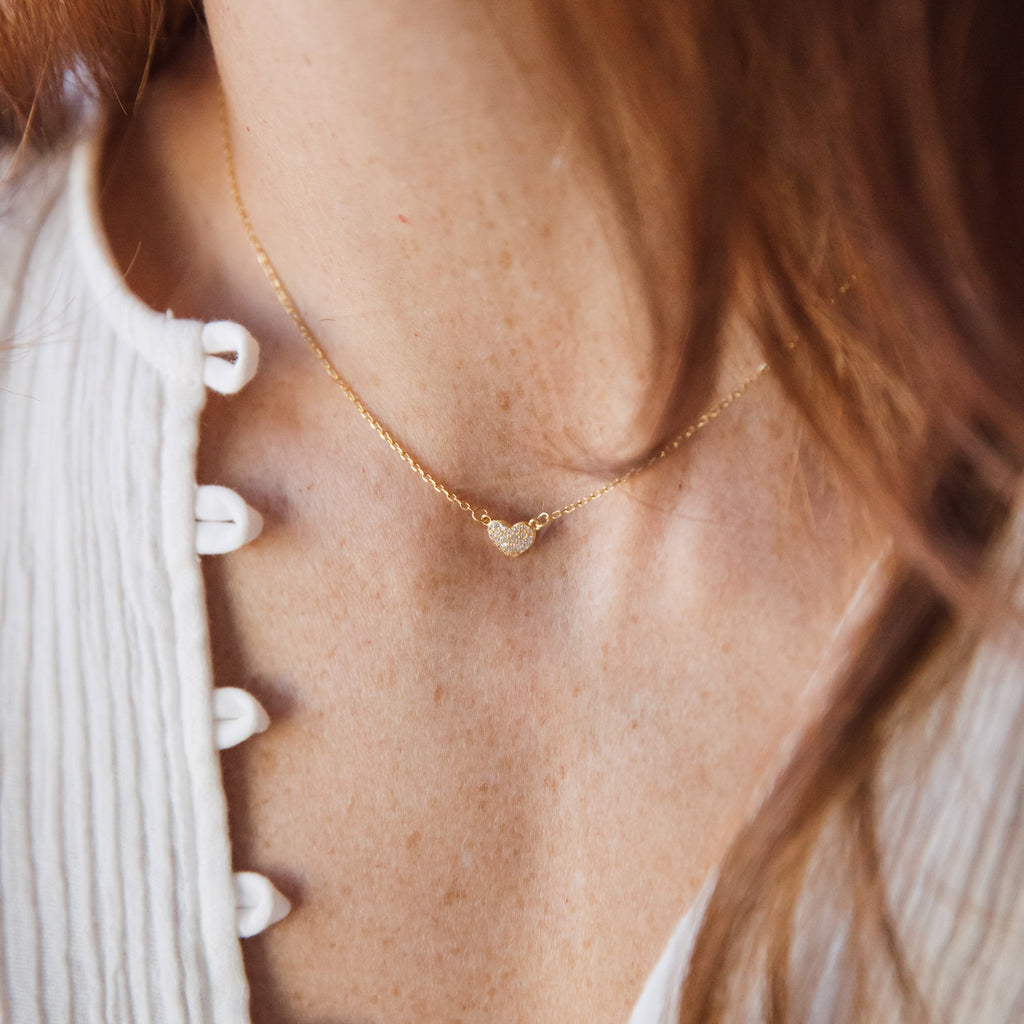 Delicate gold necklace featuring a small, pave crystal heart pendant worn close to the collarbone, styled with a textured white blouse.
