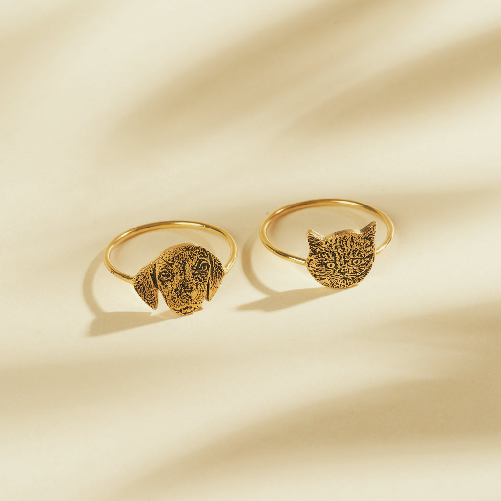 Two gold Pet Portrait Rings, each engraved with a dog or cat face, rest on a softly lit surface—these custom rings are the perfect personalized jewelry for animal lovers.