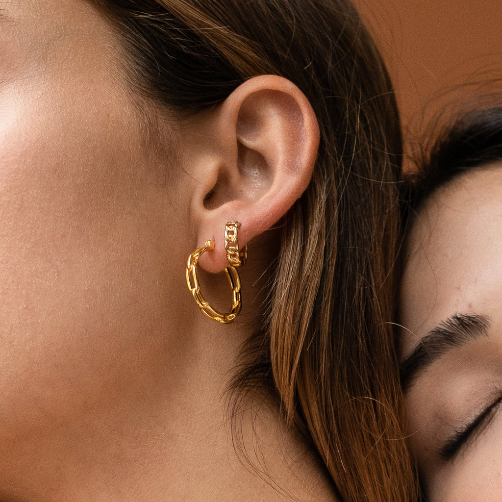Close-up of a woman's ear wearing Curb Huggies—textured gold hoop earrings with subtle chain detailing—against a brown background.