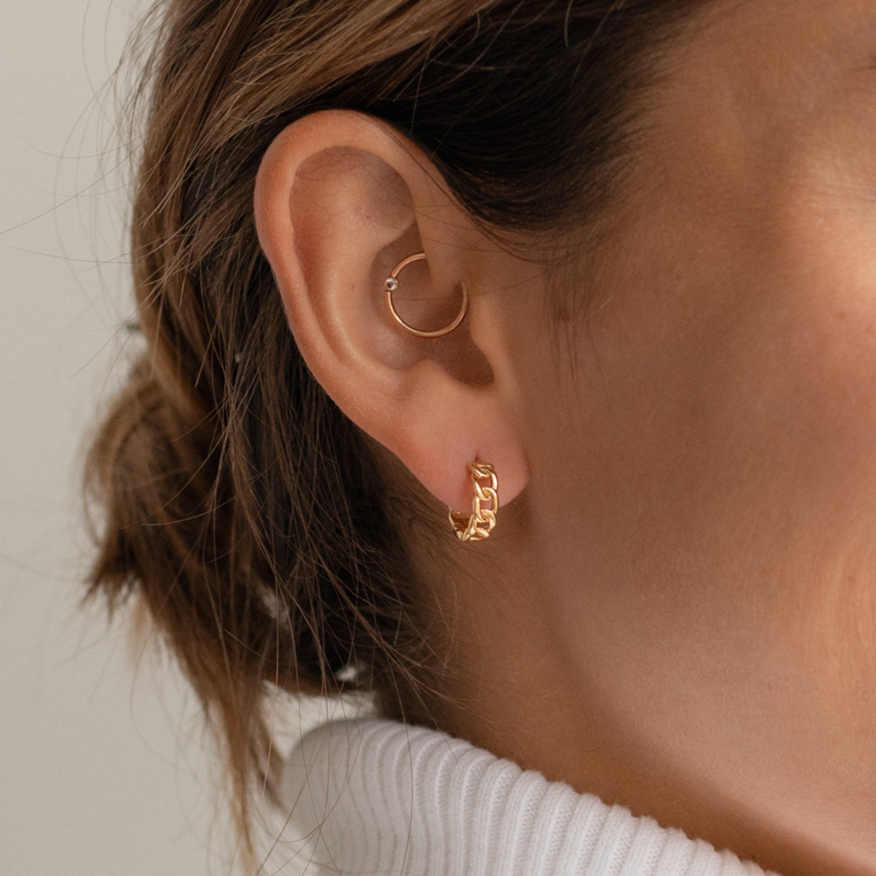 Close-up of a woman's ear wearing Curb Huggies: one gold textured hoop and one simple silver hoop, styled with a white turtleneck. These modern-luxe earrings add subtle elegance and versatile polish to any look.