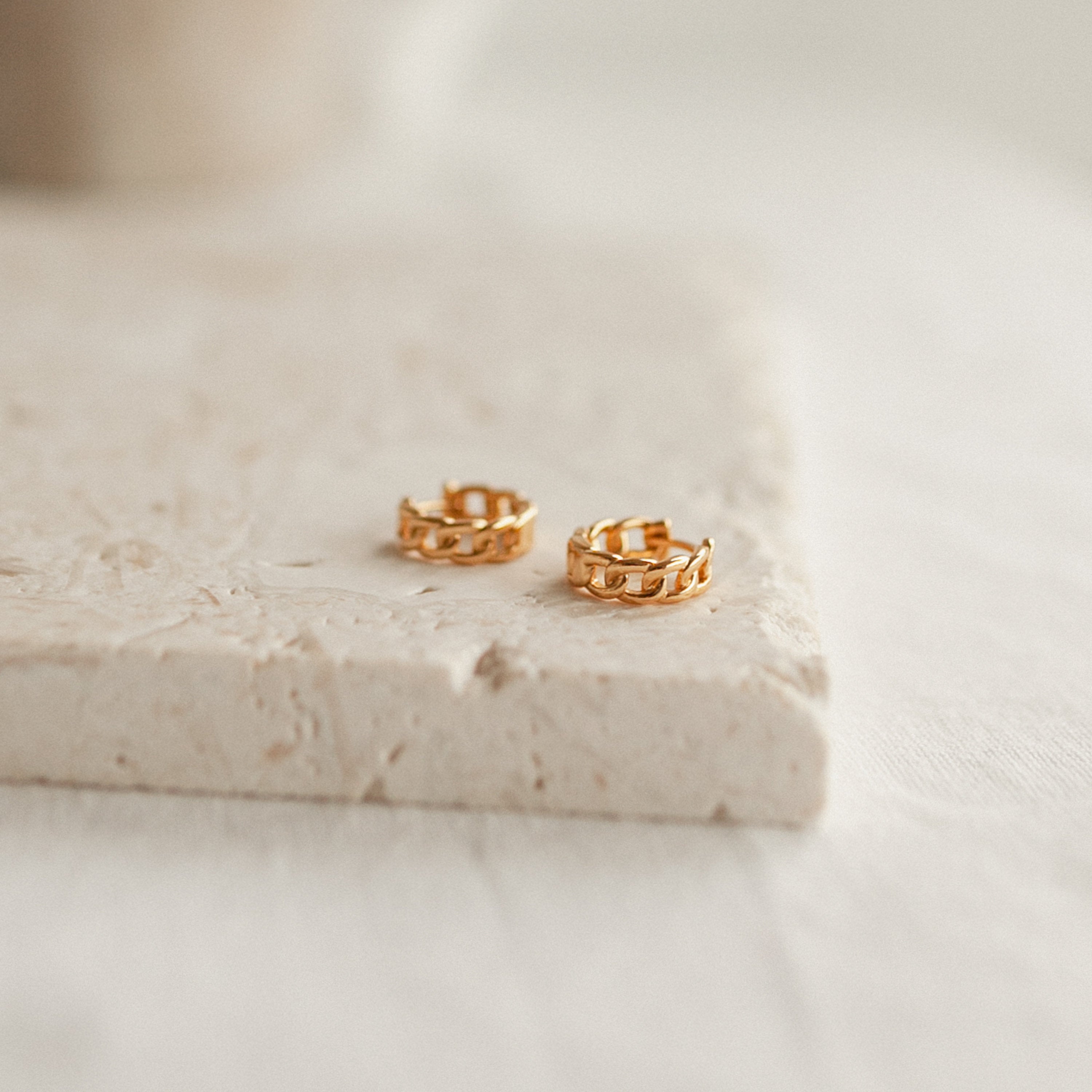 The Curb Huggies—small gold hoop earrings with chain and link detailing—rest on a light stone surface, offering an effortlessly modern-luxe look.