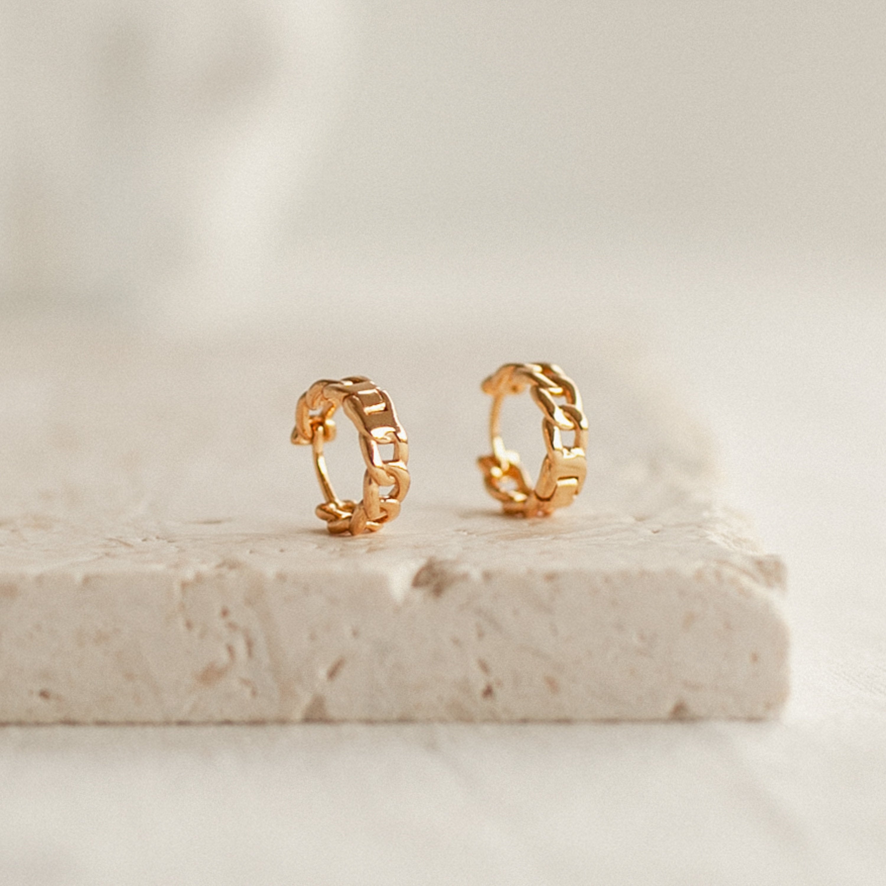 Curb Huggies gold hoop earrings feature a chunky chain design, shown on a textured, light-colored stone surface for a modern-luxe touch.