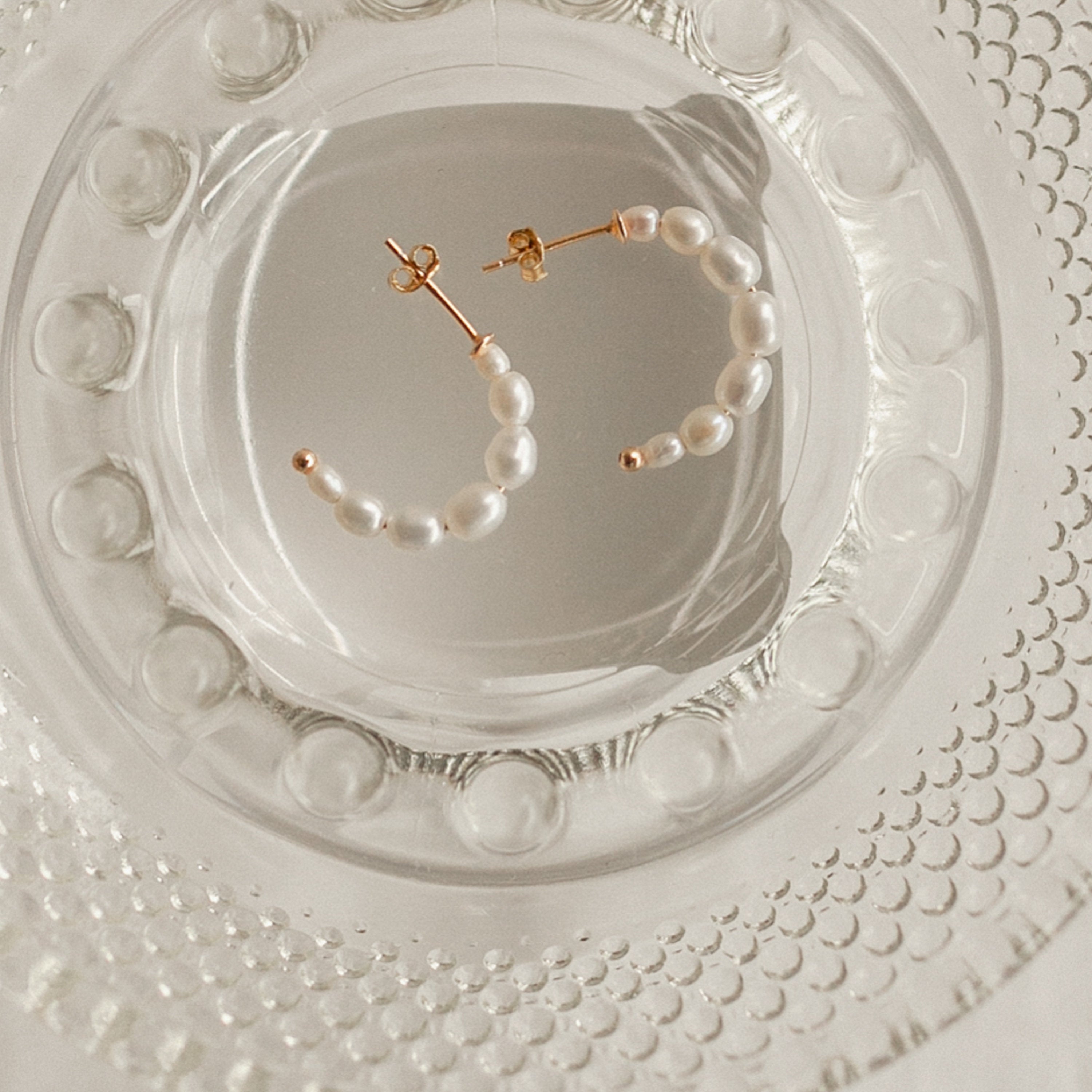 Pearl Hoops are beaded pearl earrings displayed on clear textured glass—an elegant design perfect for everyday wear.