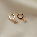 The Starburst Huggies are gold hoop earrings featuring small star-shaped pendants with pavé diamonds, shown on a soft beige fabric background.