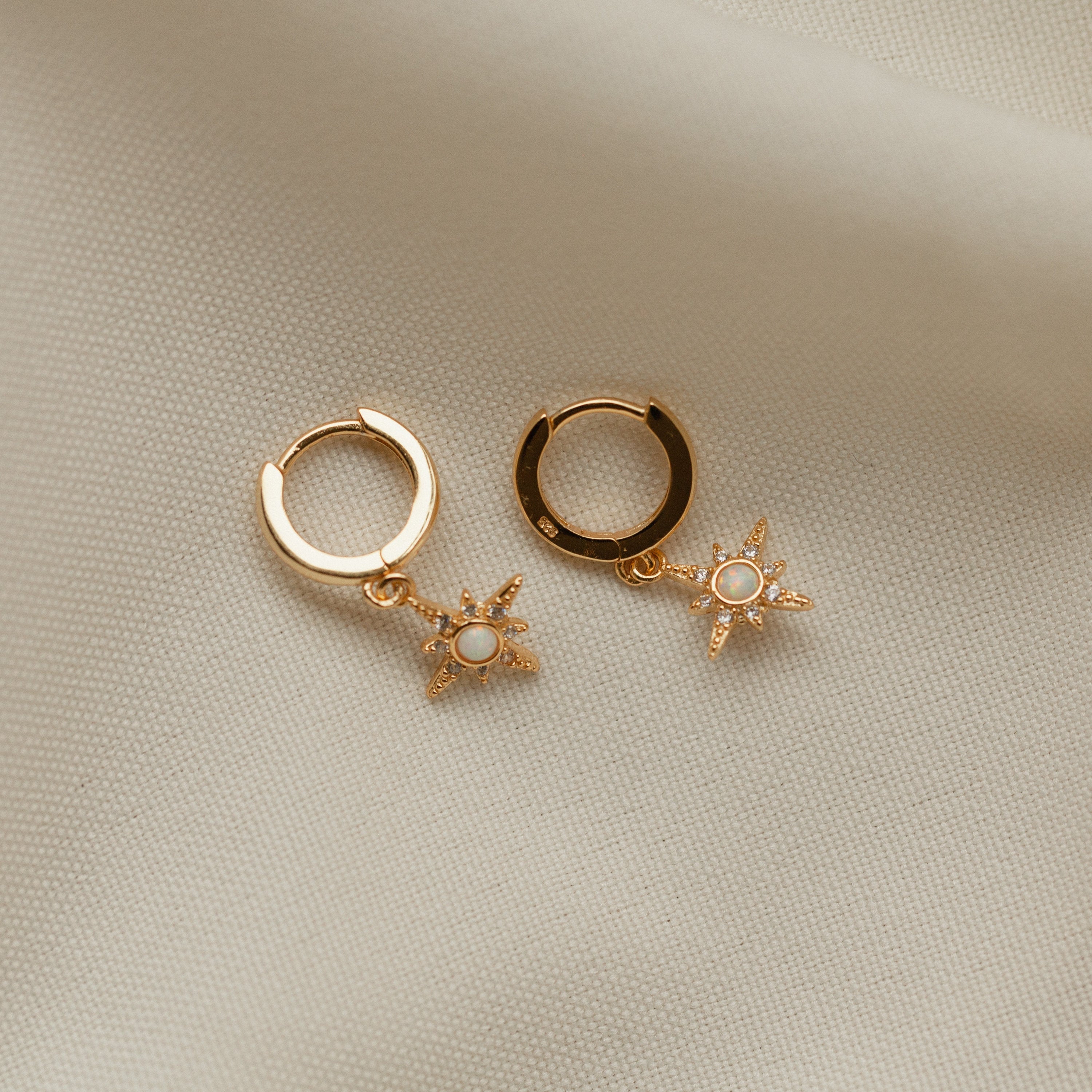 The Starburst Huggies are gold hoop earrings featuring small star-shaped pendants with pavé diamonds, shown on a soft beige fabric background.