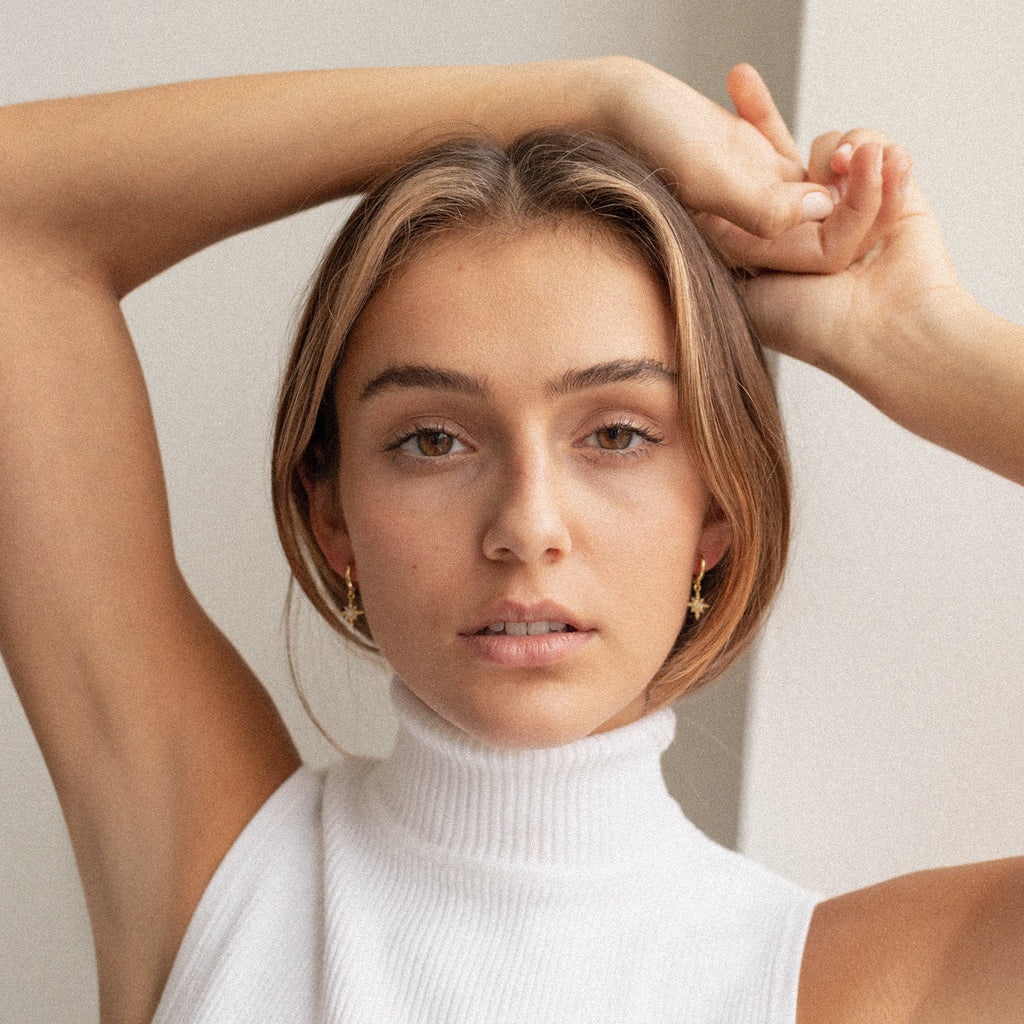 A young woman in a white sleeveless turtleneck poses with arms raised, highlighting the elegant Starburst Huggies as she looks directly at the camera.