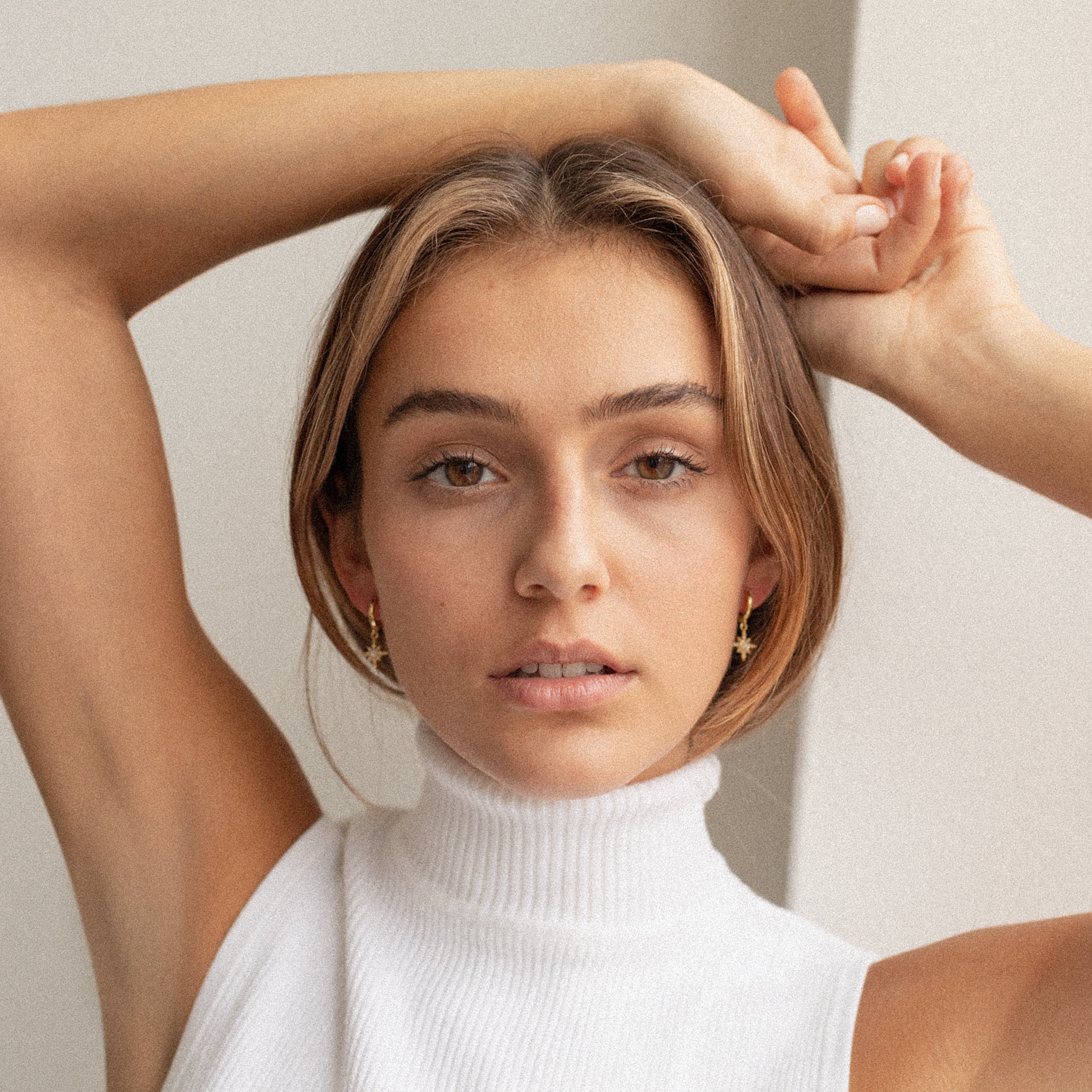 A young woman in a white sleeveless turtleneck poses with arms raised, highlighting the elegant Starburst Huggies as she looks directly at the camera.