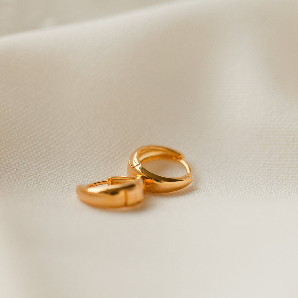 The Lucy Hinged Huggies rest on a soft, cream-colored fabric background, capturing the timeless beauty of classic hoop design.