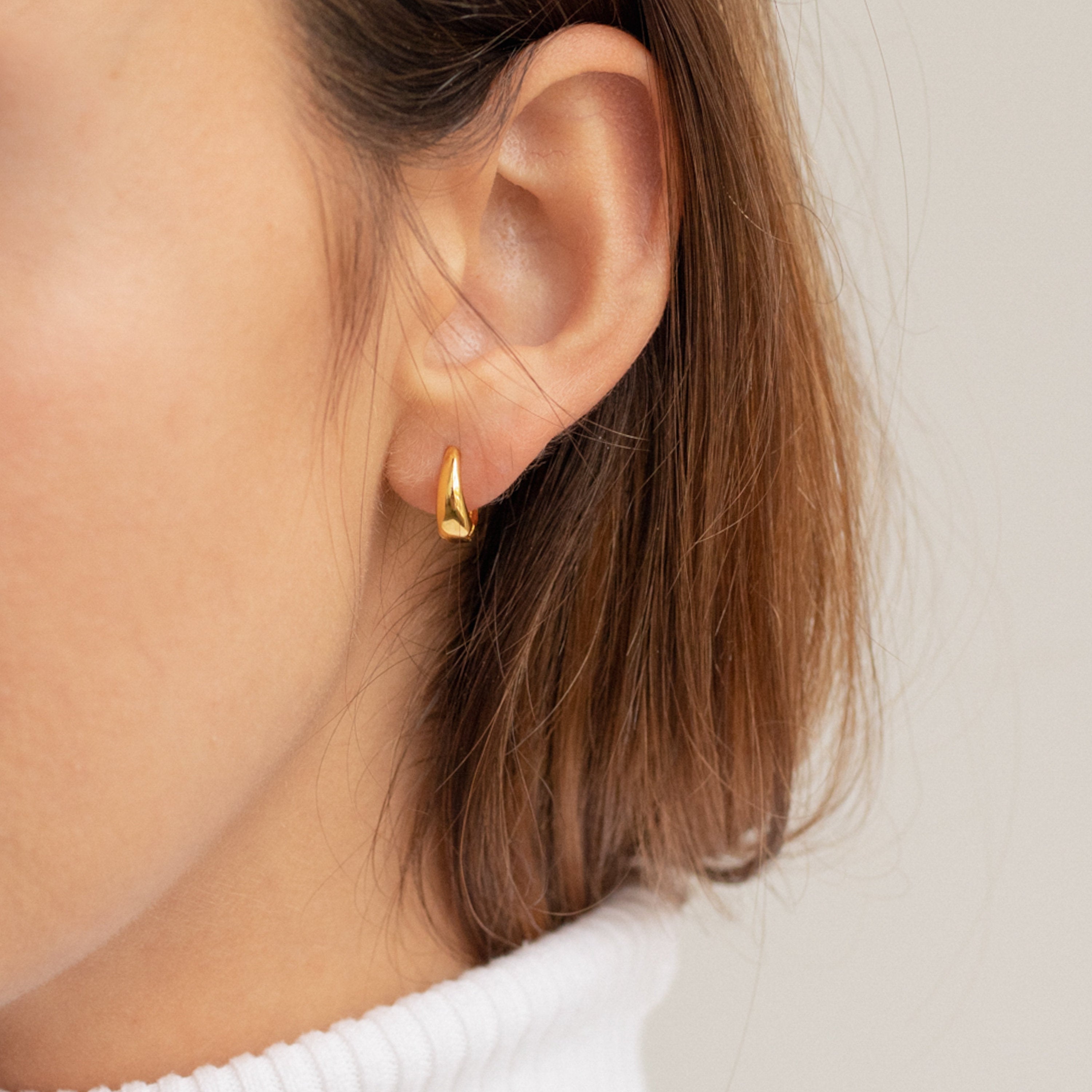 Close-up of a woman's ear adorned with the Lucy Hinged Huggies in Sterling Silver; she has short brown hair and is wearing a white top.