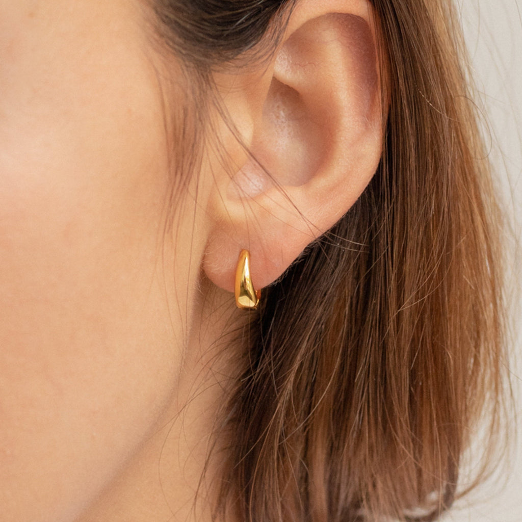 Close-up of a woman's ear wearing the gold Lucy Hinged Huggies, with short brown hair visible.