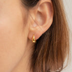 Close-up of a woman's ear wearing the gold Lucy Hinged Huggies, with short brown hair visible.