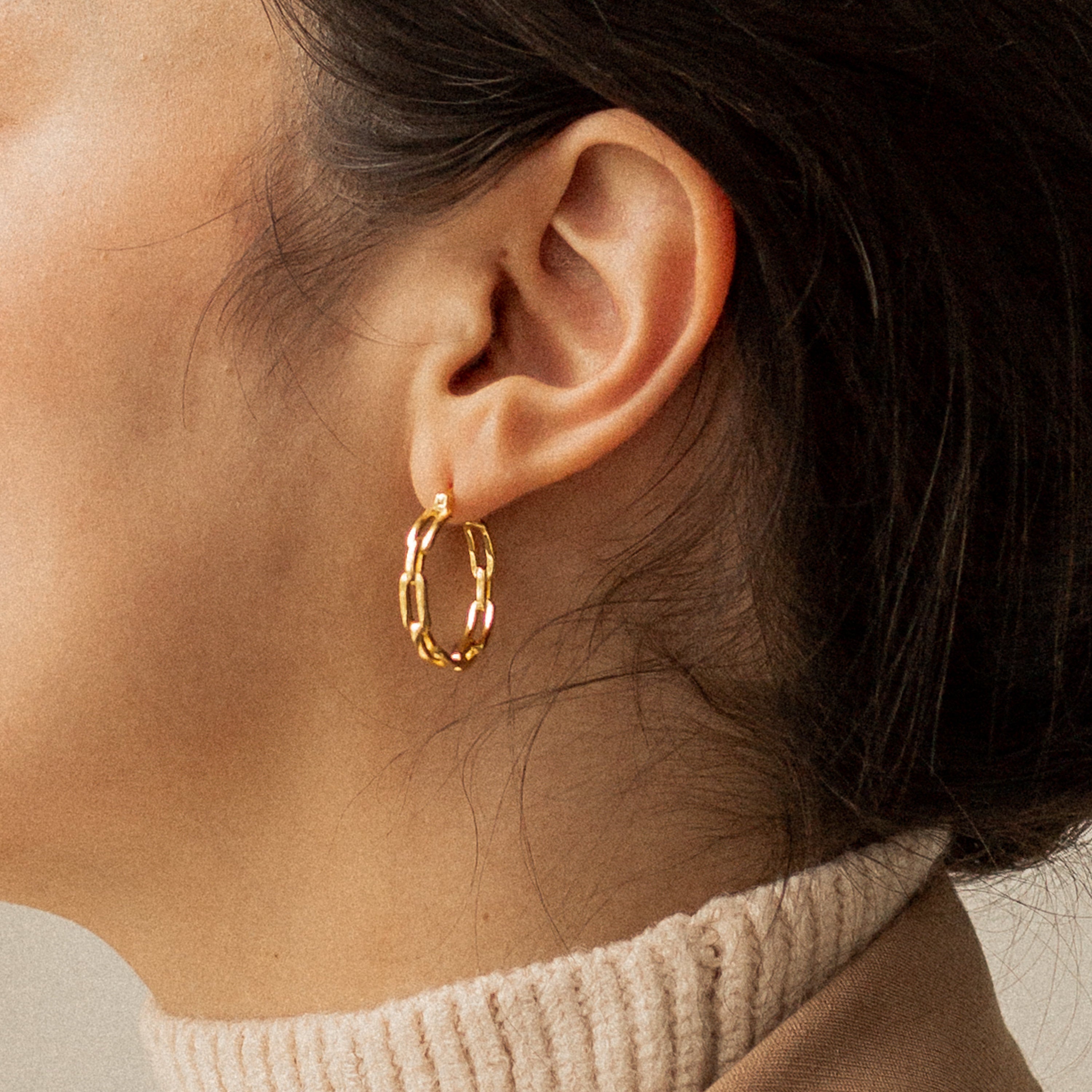 Close-up of a woman's ear adorned with Adele Link Hoops, featuring modern gold links. She has dark hair and is wearing a beige turtleneck sweater.