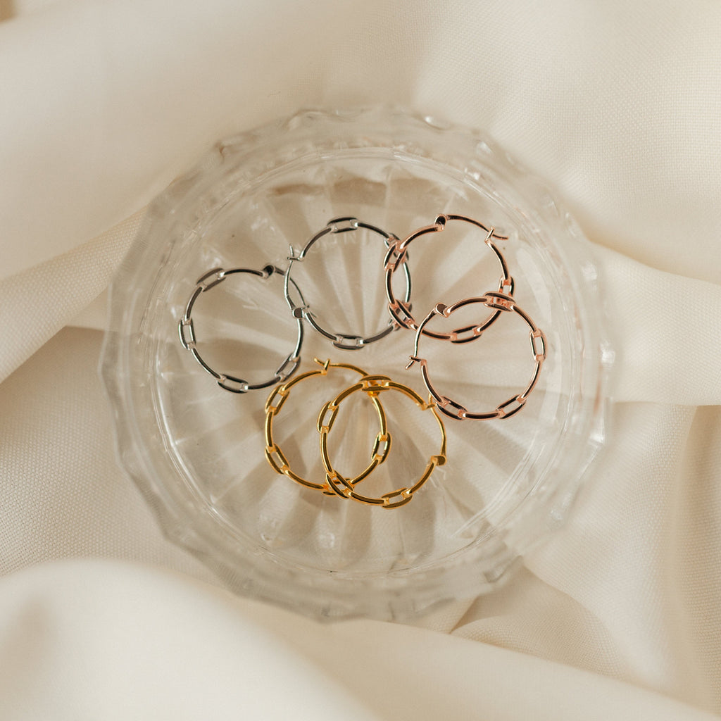 Three pairs of Adele Link Hoops—modern statement earrings with textured links in silver, rose gold, and gold—rest on a glass dish atop cream fabric.