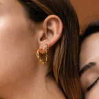 Close-up of a woman's ear wearing Adele Link Hoops—chunky gold earrings with modern, textured links—set against a blurred brown background.