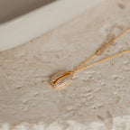 The Pave Shell Necklace features a gem-encrusted cowrie shell pendant with sparkling crystals, set on a textured beige background—an ideal accessory for a coastal cottage look.