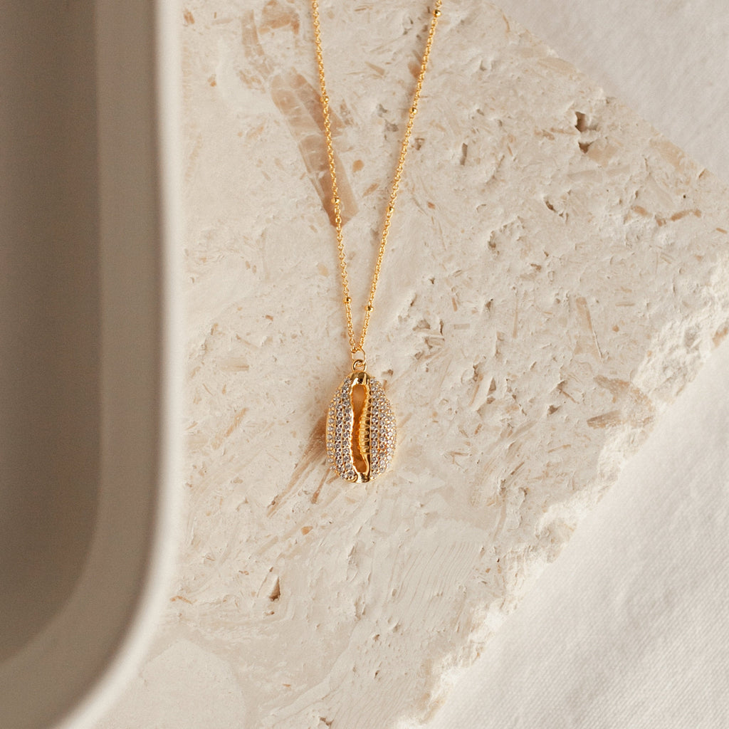 Gold cowrie shell pendant necklace with pave crystal detailing, hanging from a delicate beaded chain. displayed on a textured beige stone surface.