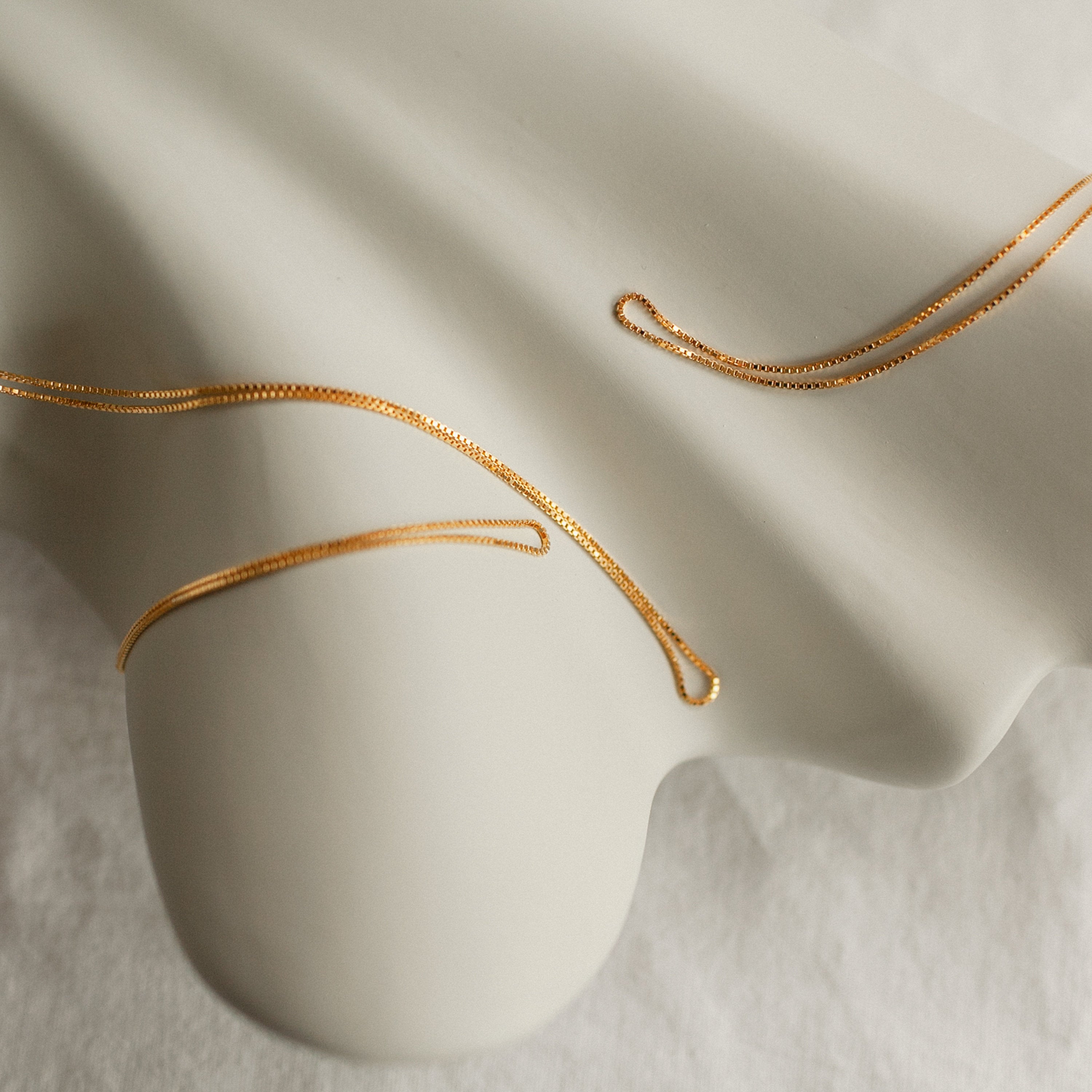 A close-up of delicate gold box chain necklaces draped artistically across an abstract cream-colored sculpture. The thin, geometric links shimmer subtly under soft lighting, emphasizing the necklace’s sleek, modern elegance and precise craftsmanship.
