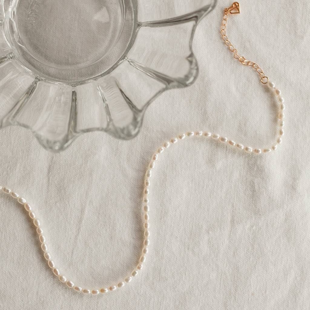 The Pearl Choker Necklace with a gold clasp is displayed next to a scalloped glass dish on a white fabric surface.