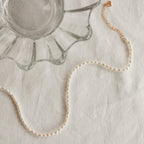The Pearl Choker Necklace with a gold clasp is displayed next to a scalloped glass dish on a white fabric surface.