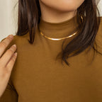 A gold herringbone necklace styled over a brown turtleneck sweater, with a hand and hair partially visible.