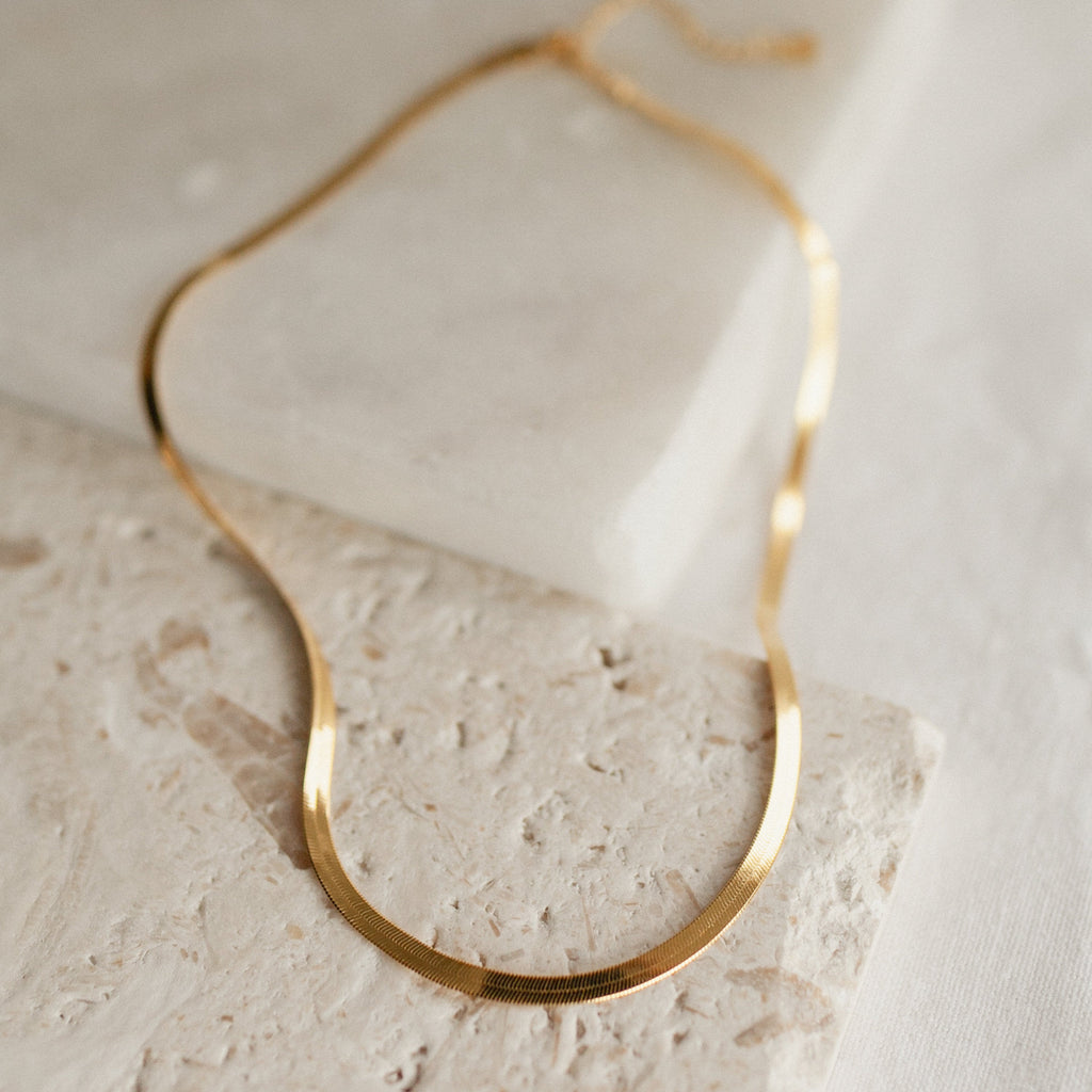A thick gold herringbone necklace laid out in an elegant curve over cream-colored textured stone.