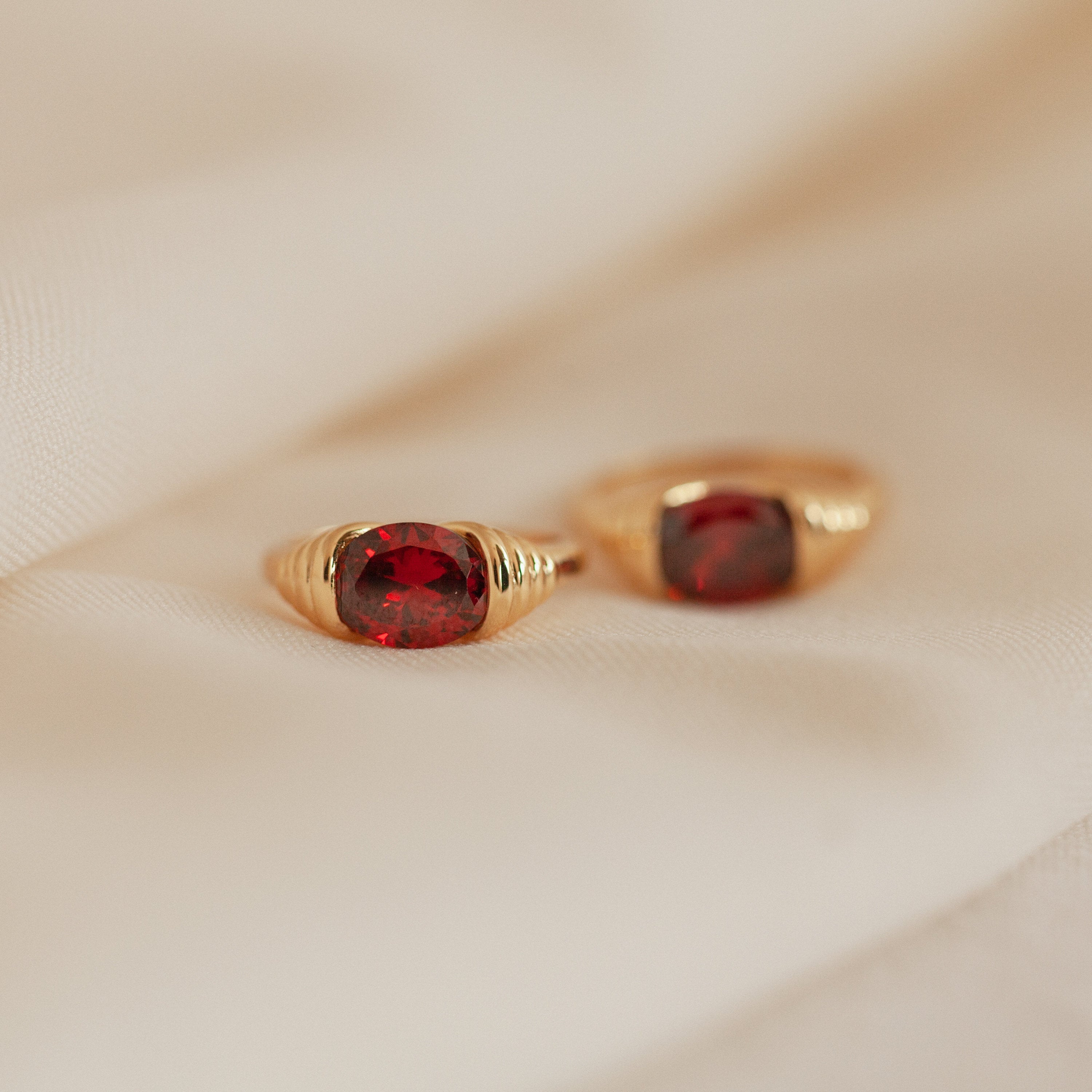 Two Garnet Signet Rings with oval red gemstones rest on soft beige fabric—a vintage-inspired pair that adds a classic touch to your signature jewelry collection.