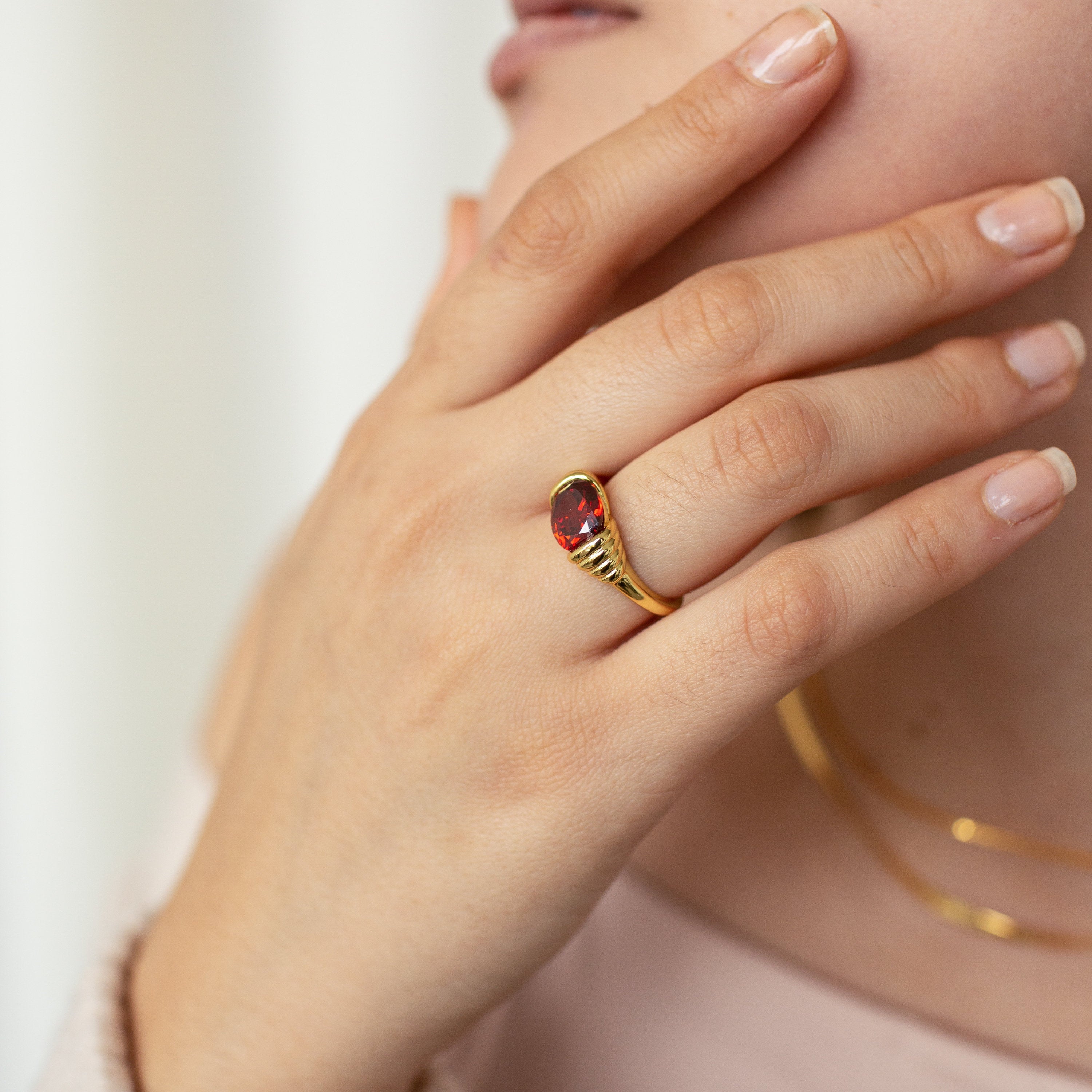 A manicured hand wears the Garnet Signet Ring, its vintage-inspired design and rich red gemstone shining as it touches the face—a striking statement piece.