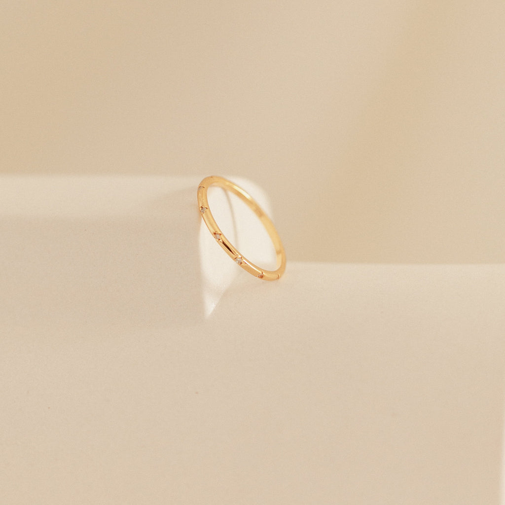 The Sprinkle Diamond Ring, ideal for stacking with other rings, is displayed on a soft beige surface with gentle light and shadow.