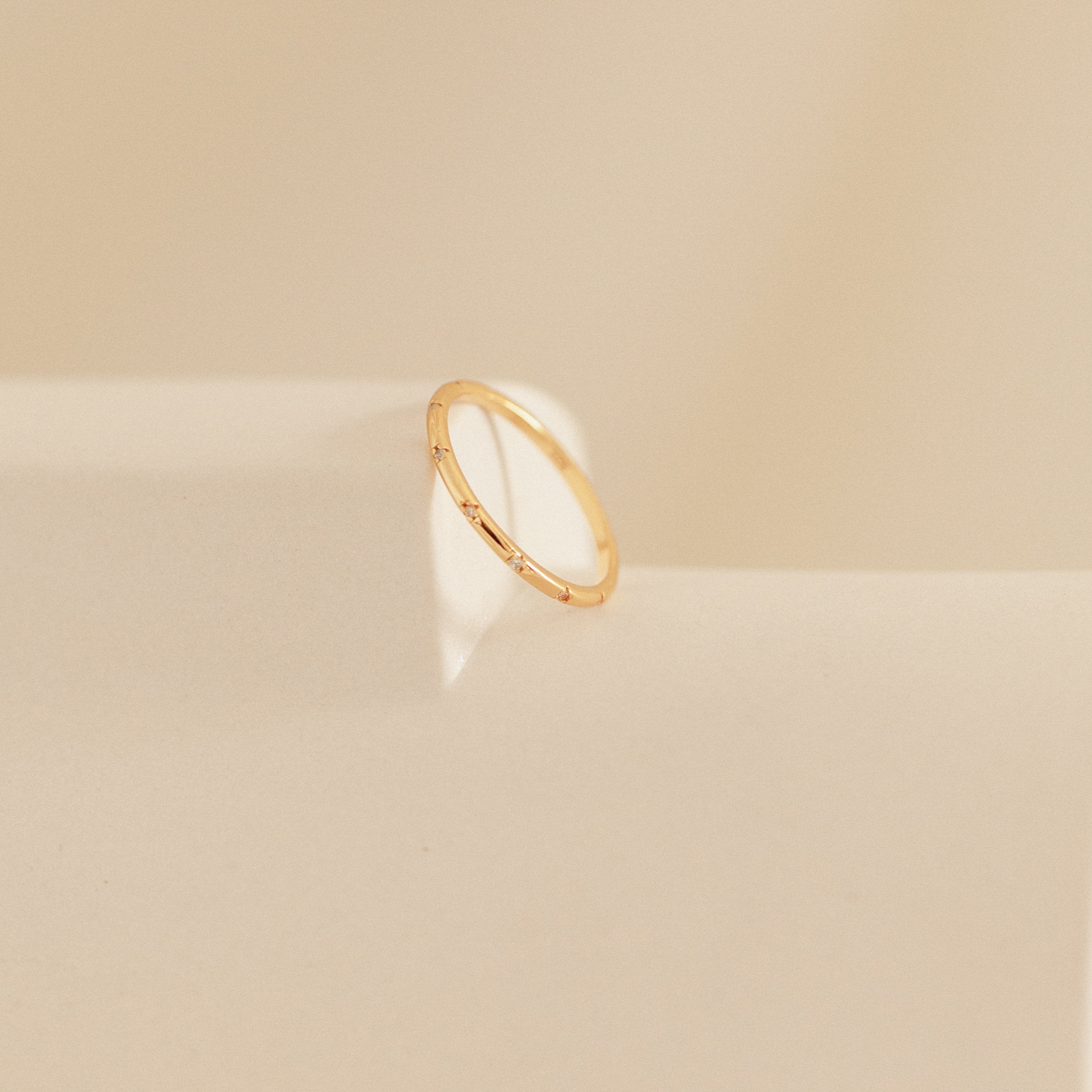 The Sprinkle Diamond Ring, ideal for stacking with other rings, is displayed on a soft beige surface with gentle light and shadow.