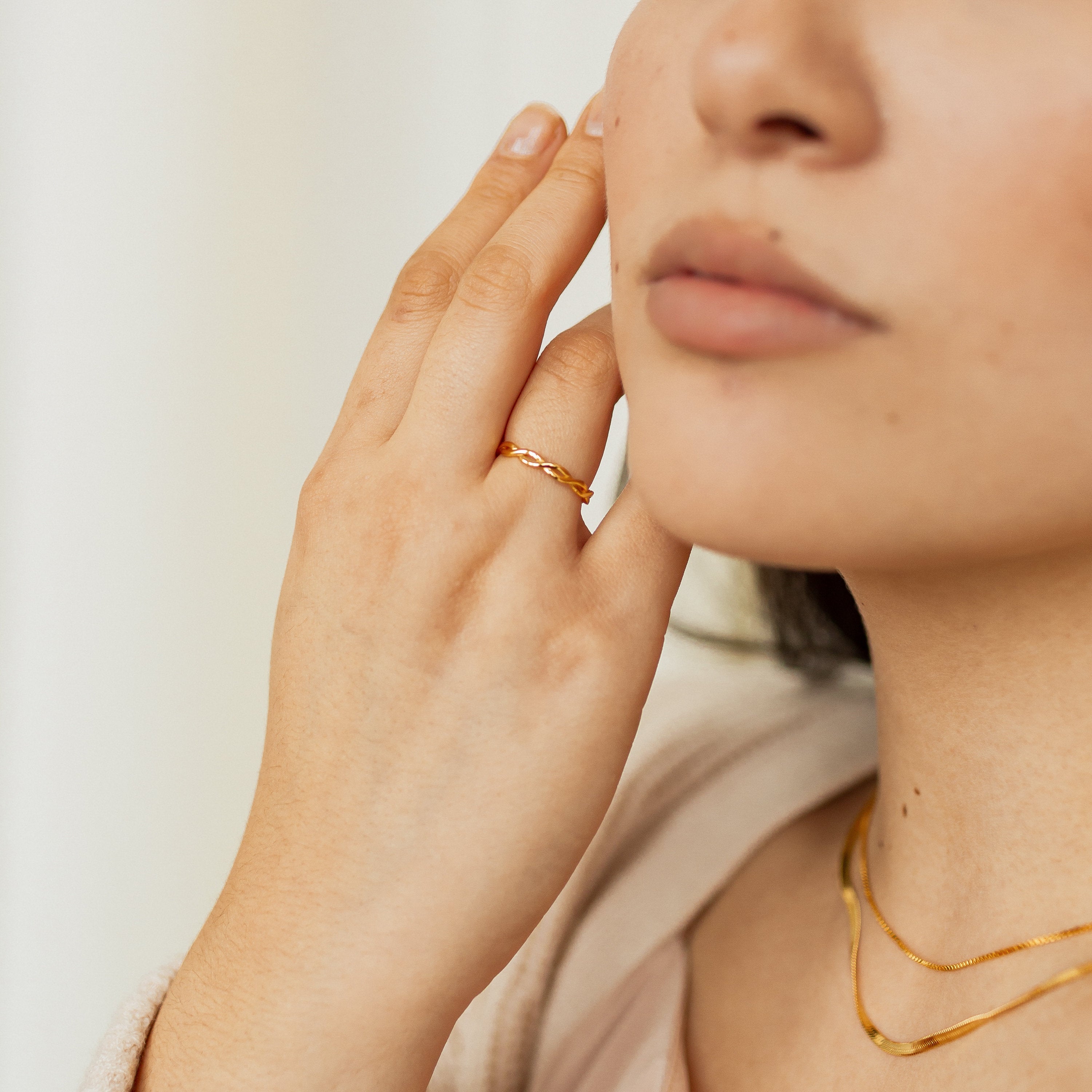 Woman touching her face, wearing the Braided Ring in gold, a sleek silver band, and layered gold necklaces; image shown from the chin down.