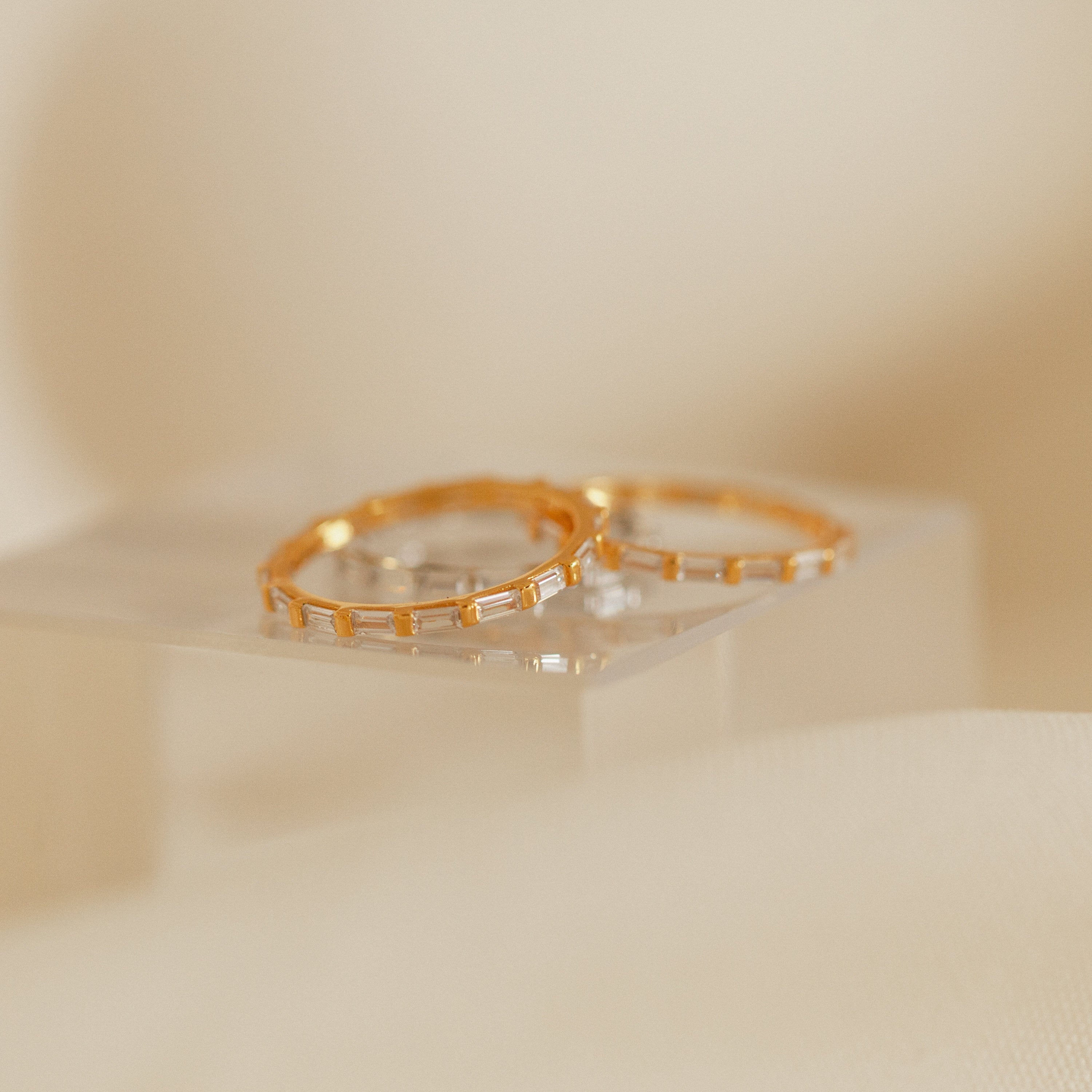 The Baguette Eternity Ring, featuring rectangular diamonds set in gold, rests on a clear square pedestal against a soft beige background, capturing modern luxe elegance.