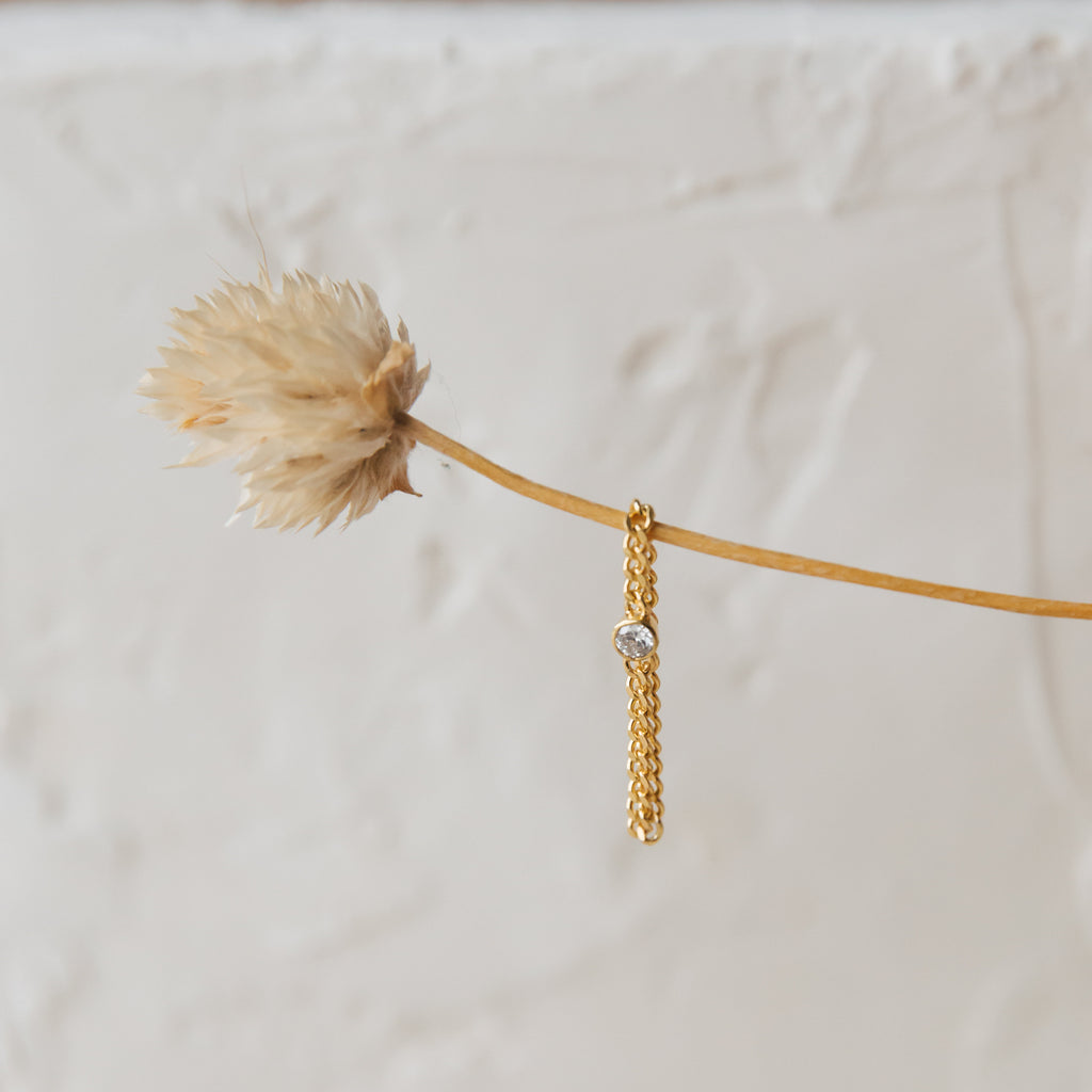 A gold chain earring with a small gemstone hangs from a dried flower stem against a textured white background, pairing perfectly with the Birthstone Curb Ring for a unique, modern style.