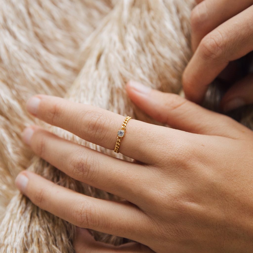 A hand wears the Birthstone Curb Ring, featuring a small personalized gemstone, resting on soft beige fabric.