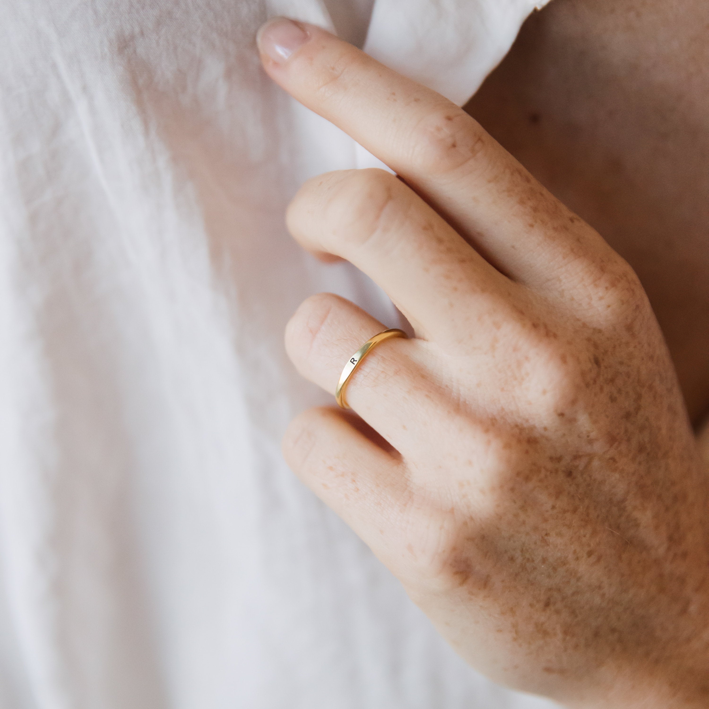 A hand with light brown freckles gently touches a white shirt, showcasing the Skinny Initial Ring as a delicate stacking piece.