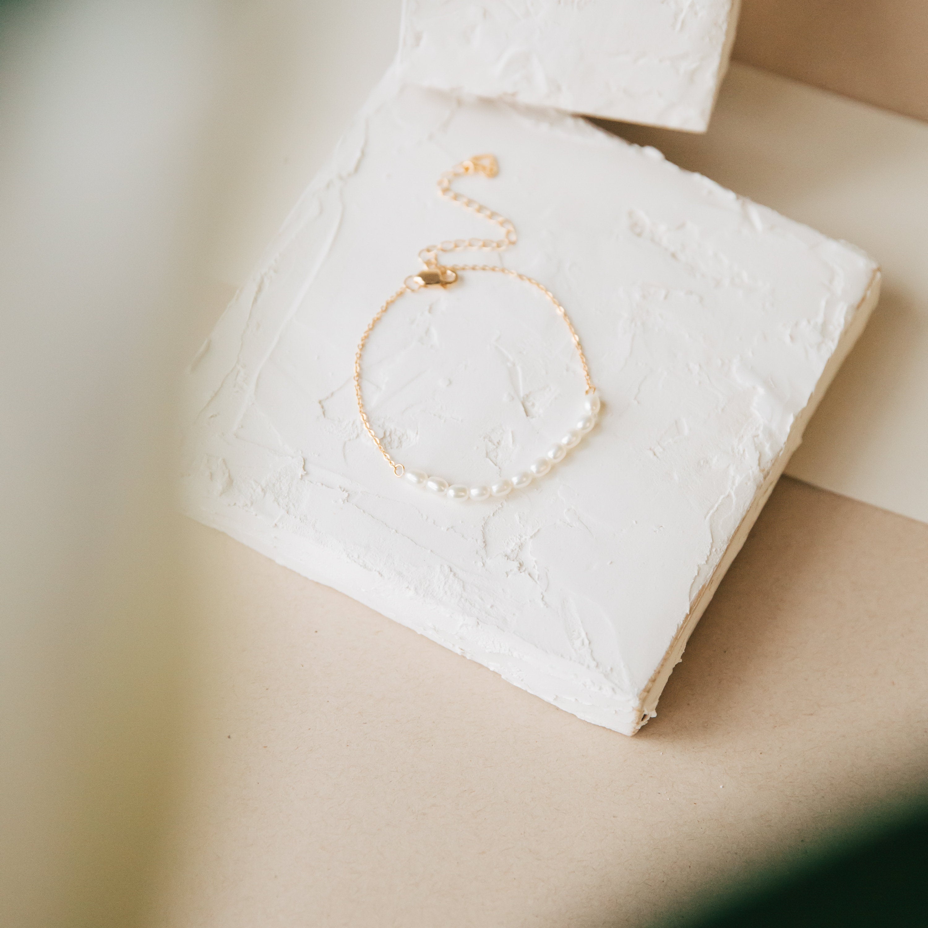 The Pearl Bead Bracelet features a gold chain with modern pearls, elegantly showcased on textured white square tiles.