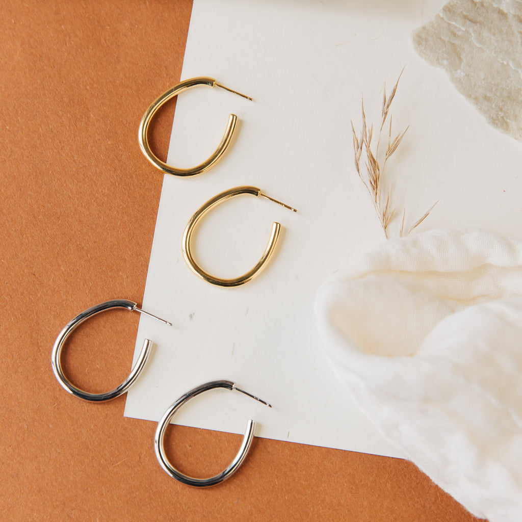 Two pairs of Anine Oval Hoops—one gold, one silver—shown on brown and white surfaces. These stylish oval hoop earrings elevate any look.
