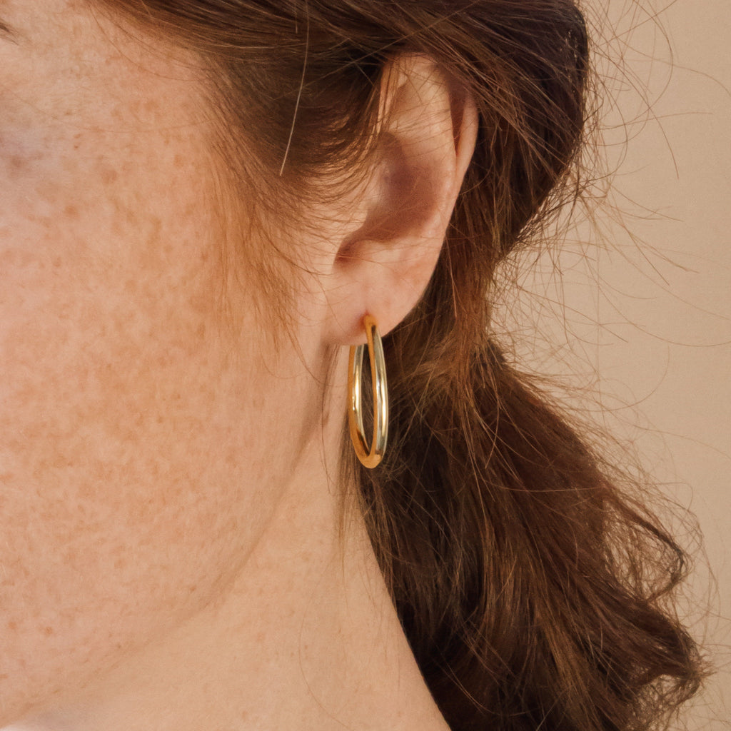 Close-up of a person with freckles and brown hair tied back, wearing Anine Oval Hoops.