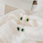Two pairs of green Emerald Drop Earrings—one gold teardrop and one silver teardrop style—are displayed on soft white fabric.