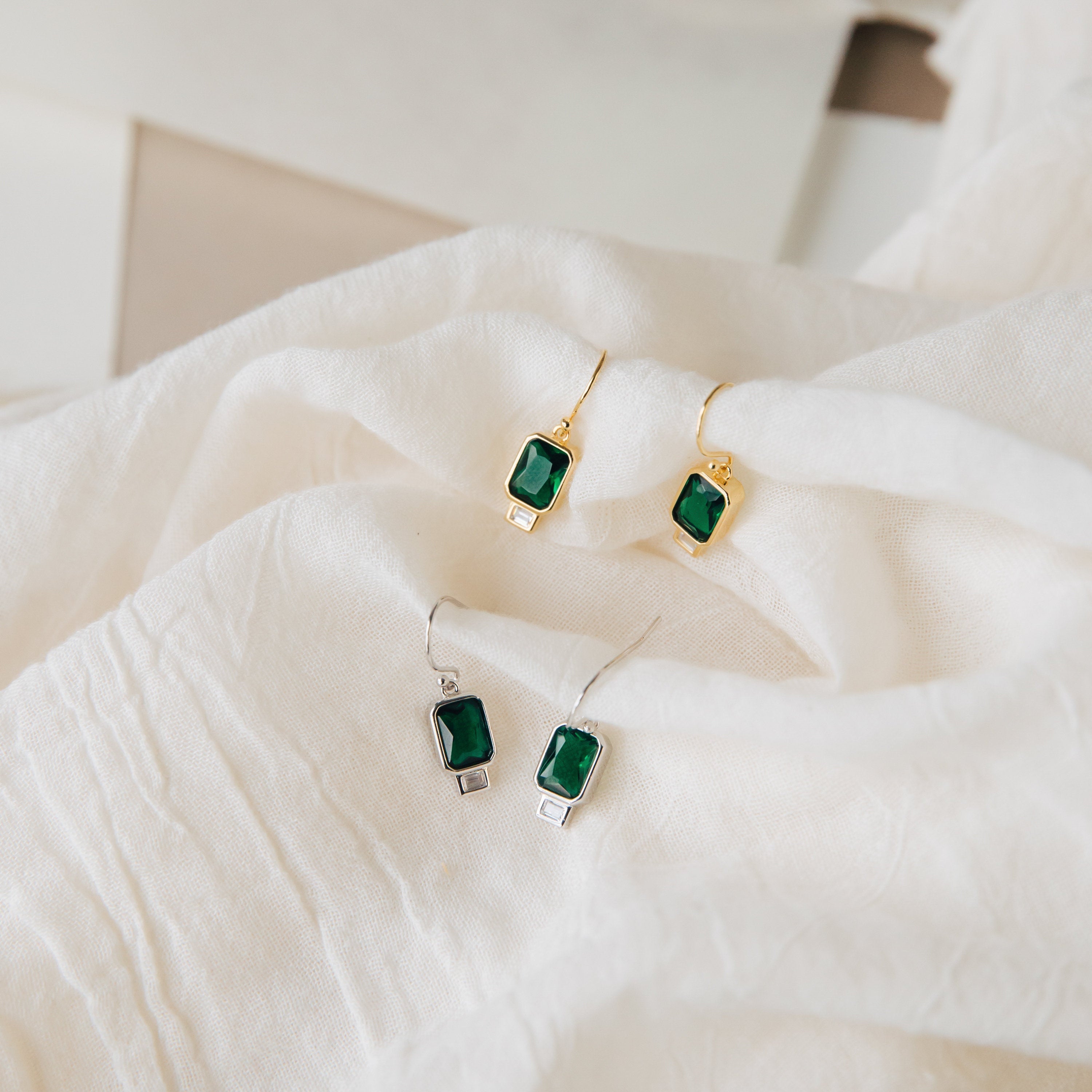 Two pairs of green Emerald Drop Earrings—one gold teardrop and one silver teardrop style—are displayed on soft white fabric.